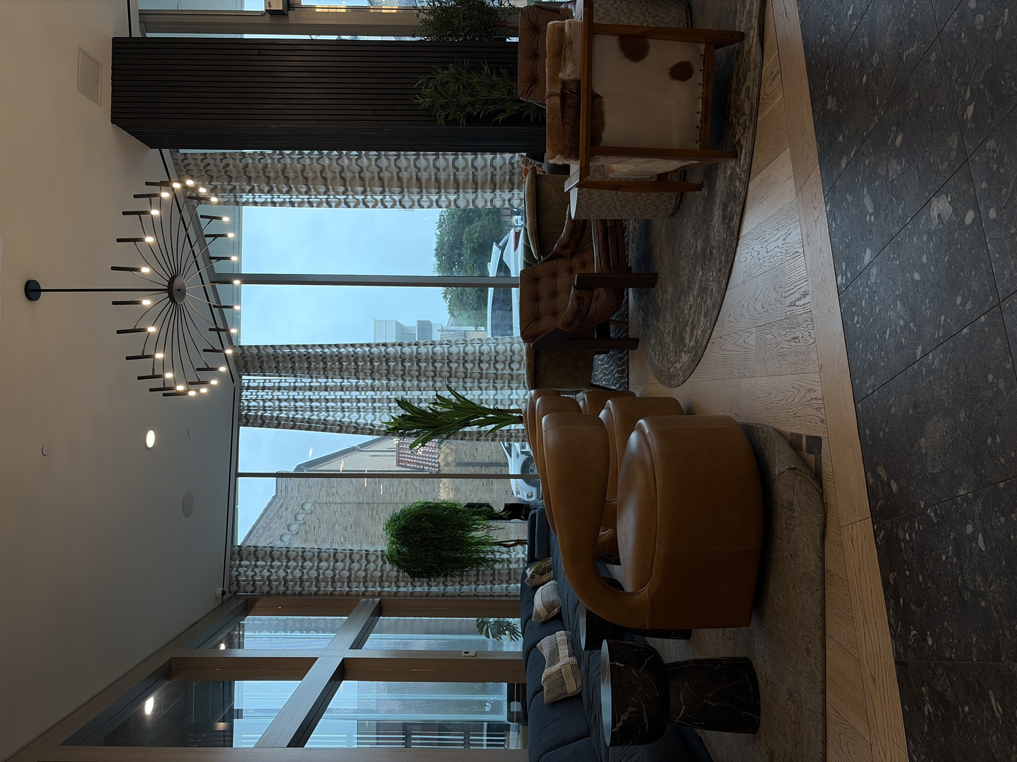 This media file captures a modern and sophisticated lobby or lounge area, likely in Austin, United States of America. No people are visible, presenting a serene and unoccupied space.
The scene features a diverse arrangement of comfortable seating. To the left, a long dark grey fabric sofa is adorned with several textured throw pillows. Adjacent to it are two circular side tables with a black marble-like finish. Further into the room, several brown leather armchairs with rounded backs are situated on a large, irregularly shaped grey rug. Another distinct armchair, upholstered in brown tufted leather with wooden arms, is visible. Two more armchairs, featuring natural wooden frames and speckled cowhide or fur upholstery, are positioned on the far right.
Architectural elements include expansive floor-to-ceiling windows that provide abundant natural light and a view of the outdoors. These windows are framed by patterned, light-toned curtains. A striking black, wagon-wheel-style chandelier with numerous small lights hangs from the white ceiling, which also features recessed lighting and a rectangular vent. A dark wooden slatted accent wall adds texture to the space.
Outside the windows, the sky appears overcast or gloomy, suggesting a cloudy day. A street is visible, along with a white vehicle, a mature tree, and a brick building with a distinctive facade.
The flooring transitions from light-toned wooden planks in the center to dark, speckled square tiles on the right. Decorative potted plants, including tall, leafy varieties and a bushy green plant, are strategically placed throughout the area, enhancing the natural aesthetic. The word "spkr" is faintly visible on a white ceiling panel in one of the cropped images, indicating a speaker. The overall ambiance is one of contemporary elegance and comfort.