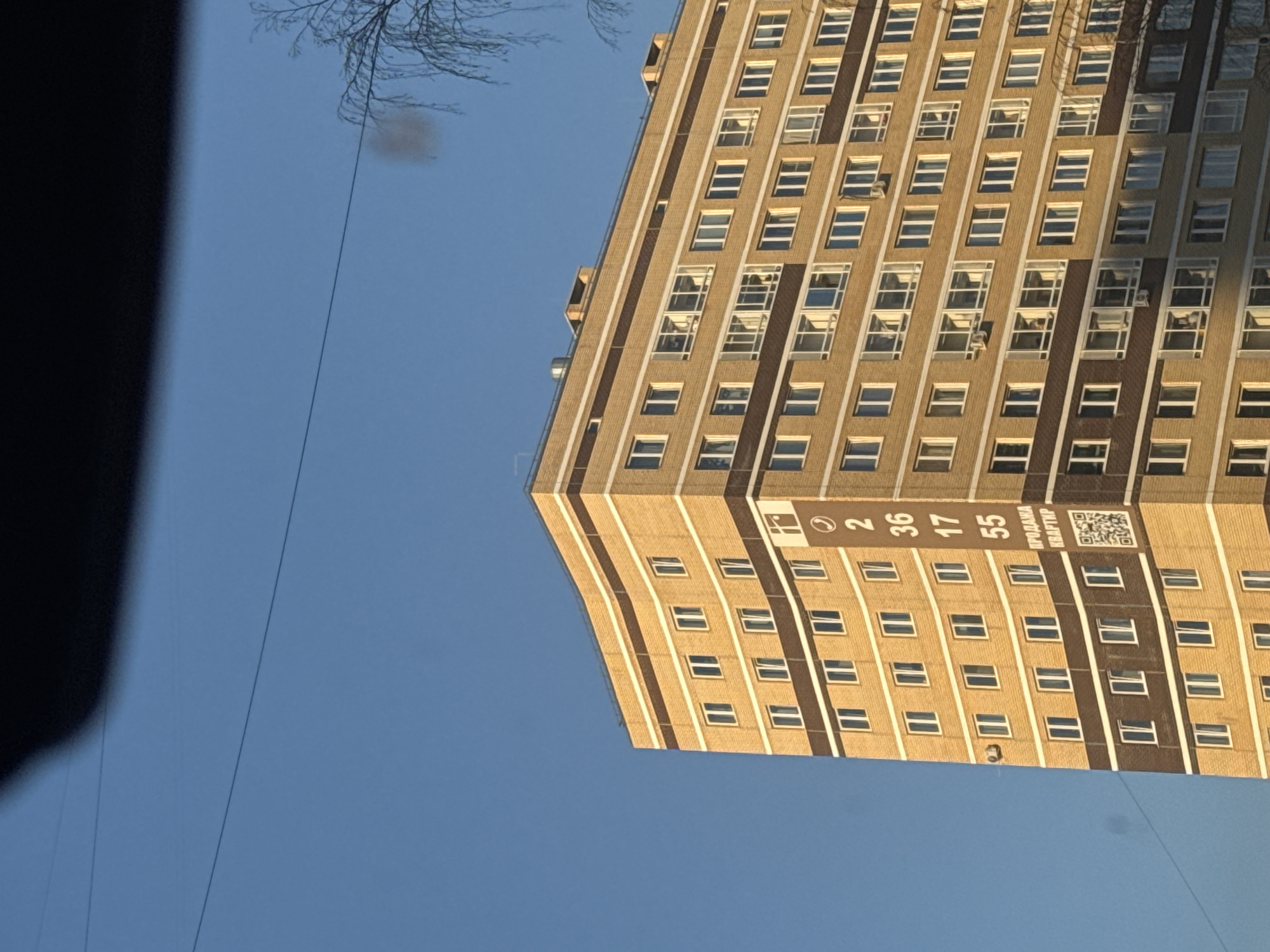 The image depicts a tall, multi-story building in Perm, Russia. The building is constructed of light brown brick and has numerous windows arranged in a grid-like pattern. A banner is affixed to the side of the building, displaying numbers and Cyrillic text, including "ПРОДАЖА КВАРТИР," which translates to "Apartment Sale." A QR code is also present on the banner. The sky is clear and blue, and bare tree branches are visible at the top left of the frame. The image is taken from a low angle, looking upwards towards the building. A dark, undefined object is in the left foreground.