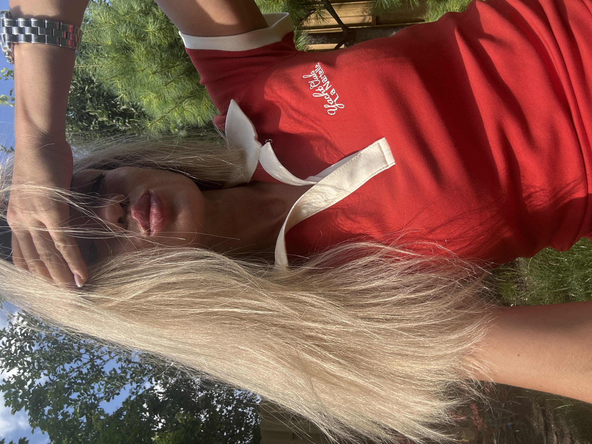 An outdoor medium shot captures a fair-skinned woman with long, blonde hair and a red collared shirt. She is holding her right hand up to her forehead, shielding her eyes. Her lips are pursed as if blowing a kiss. A silver watch is visible on her left wrist. The background consists of green trees and a bright blue sky. The image does not contain any information about the city of Armavir, Russia.