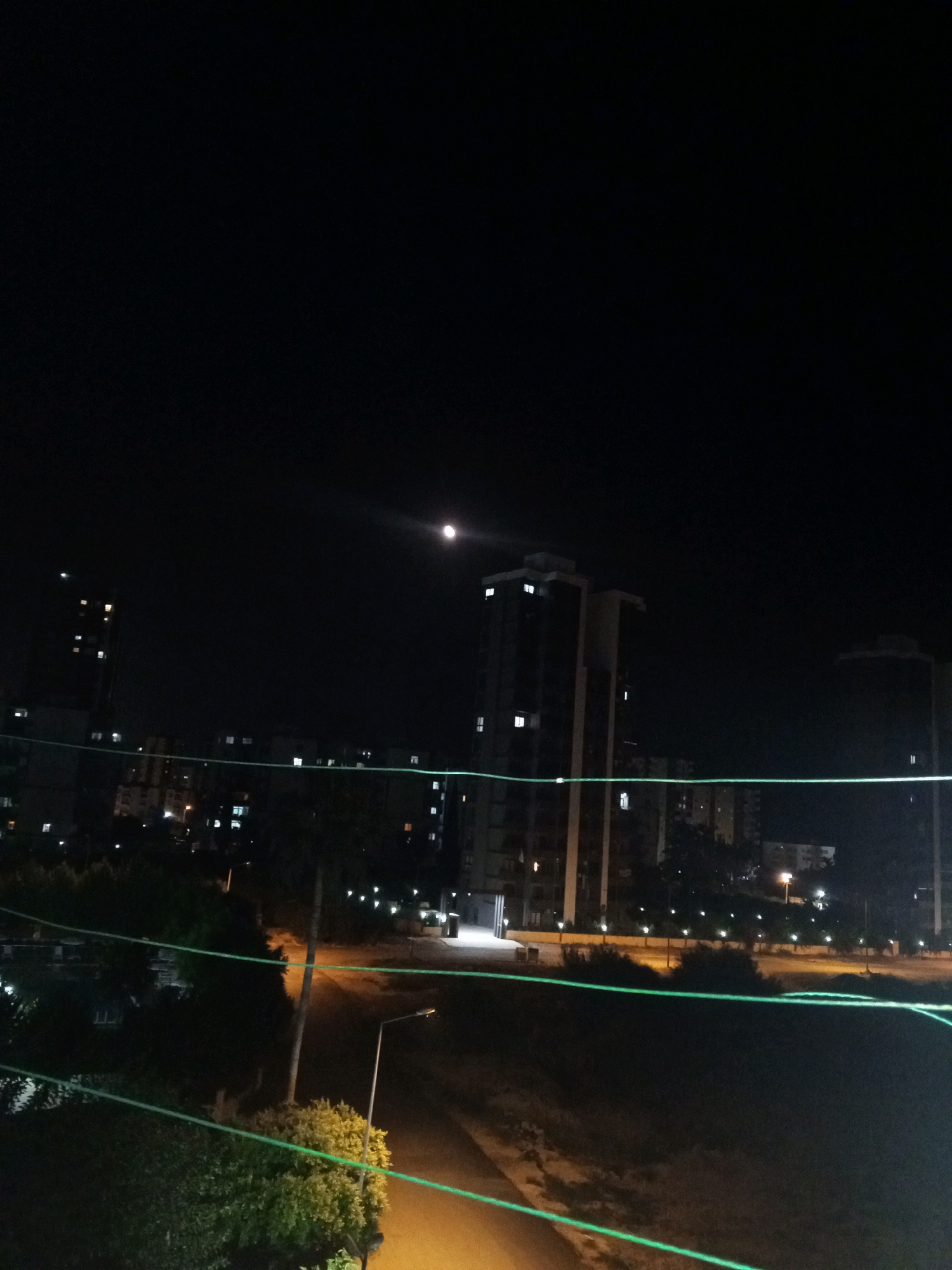 The image is a nighttime, long-exposure photograph taken from a high vantage point, likely a balcony or rooftop, in Mezitli, Turkey. The dark sky is illuminated by the moon, which is visible in the upper right quadrant, emitting a bright light. In the mid-ground, several multi-story apartment buildings are lit, with numerous windows showing interior lights. The foreground is dominated by dark foliage and the outline of a structure, possibly a building or fence. Two bright green lines, possibly from laser pointers or light displays, cut across the scene horizontally in the lower half of the image, creating a visual disruption. The overall impression is a cityscape at night with artificial lighting from buildings and the moon.