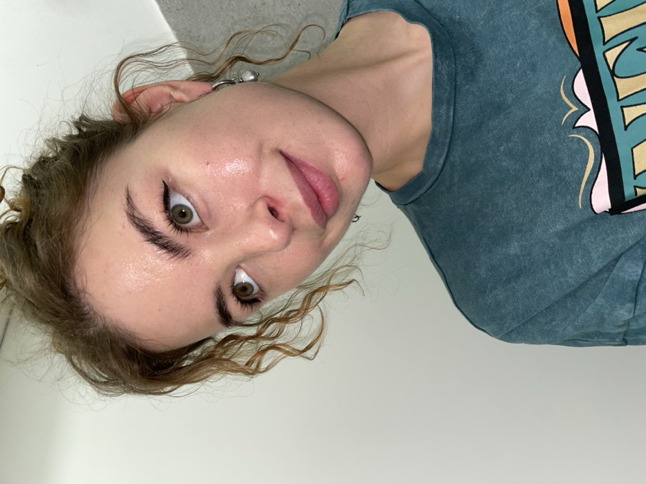 This image presents a close-up, slightly angled portrait of an adult female, situated in Budva, Montenegro. The subject exhibits fair, dewy skin, light brown curly hair gathered in an updo, and prominent green eyes. Black eyeliner is visible along the upper lash line, and her eyebrows are full. Her expression involves a subtle smile and an upward gaze directed towards the upper right of the frame. A single silver-toned hoop earring, featuring a white, irregular pearl drop, is present on her visible right ear. She is attired in a teal-colored, short-sleeved, crew-neck t-shirt, part of a faded graphic design is visible on the chest area. The background is a plain, light-colored, possibly painted, interior wall, providing a neutral setting. The scene is illuminated by soft, direct lighting.