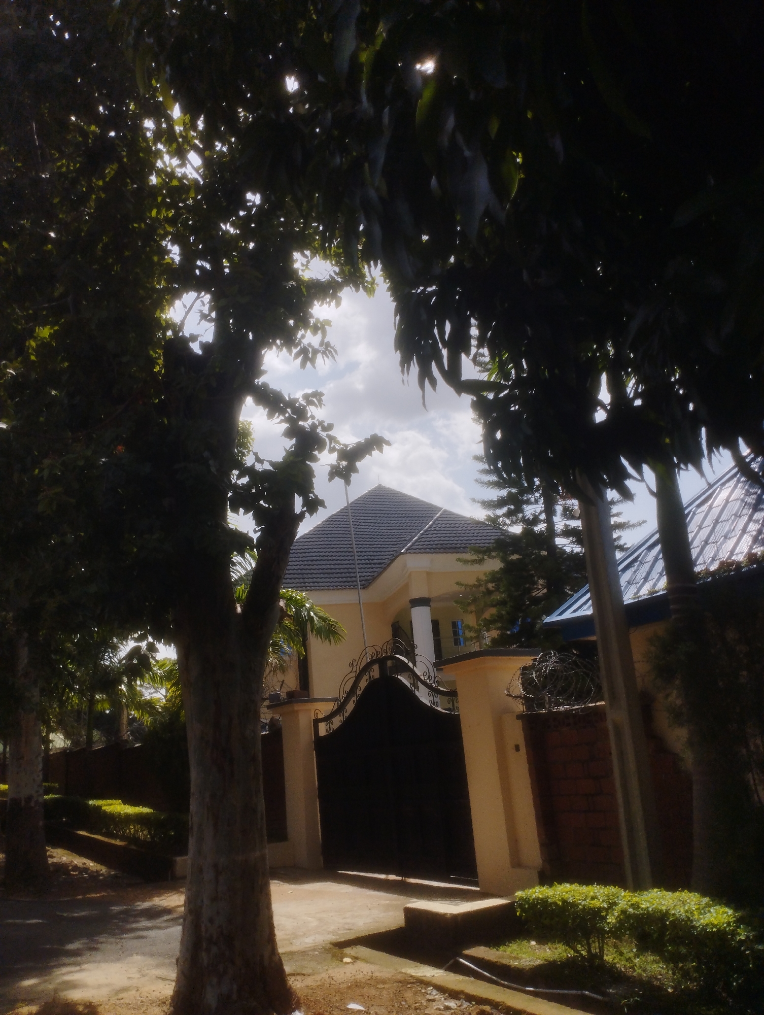 The image shows a street view with a large tree in the foreground. Behind the tree, a modern house with a dark tiled roof is visible, featuring a large black gate with ornate metalwork and a tall brick wall topped with barbed wire. Sunlight filters through the leaves of the trees, creating dappled light on the ground. The scene appears to be a residential area in Jos, Nigeria.