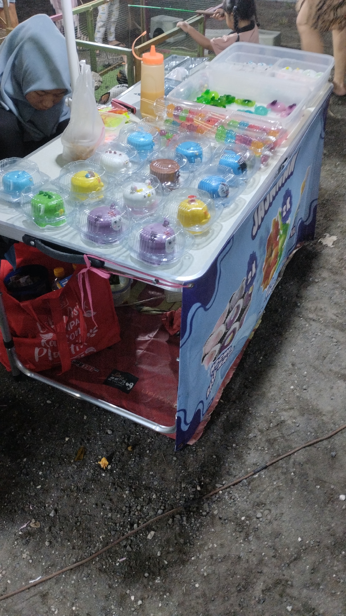 The image shows a street vendor stall in Sambungrejo, Indonesia, likely at dusk or in the evening given the lighting. The stall is set up on a rough, gravelly surface. The primary focus is a table laden with colorful food items, likely desserts or candies, arranged in individual plastic containers.
A woman wearing a light blue hijab is visible on the left side of the frame, hunched over the table and appearing to be engaged with the items. To her right, a child with braided hair is visible in the background, actively reaching towards something, possibly interacting with the stall or a game.
The table itself is covered with a blue and white patterned cloth or banner. Visible text on this banner appears to be in Indonesian and includes "JAJAMAN" along with cartoonish illustrations of food items, possibly sushi. There are also various containers of colorful candies or jellies on the table, as well as a plastic squeeze bottle filled with an orange liquid, likely a syrup or sauce.
Beneath the table, a red tote bag is visible, along with other items stored there. The overall scene suggests a lively, possibly festive, outdoor environment, common at local markets or street gatherings in Indonesia. The atmosphere seems casual and community-oriented.