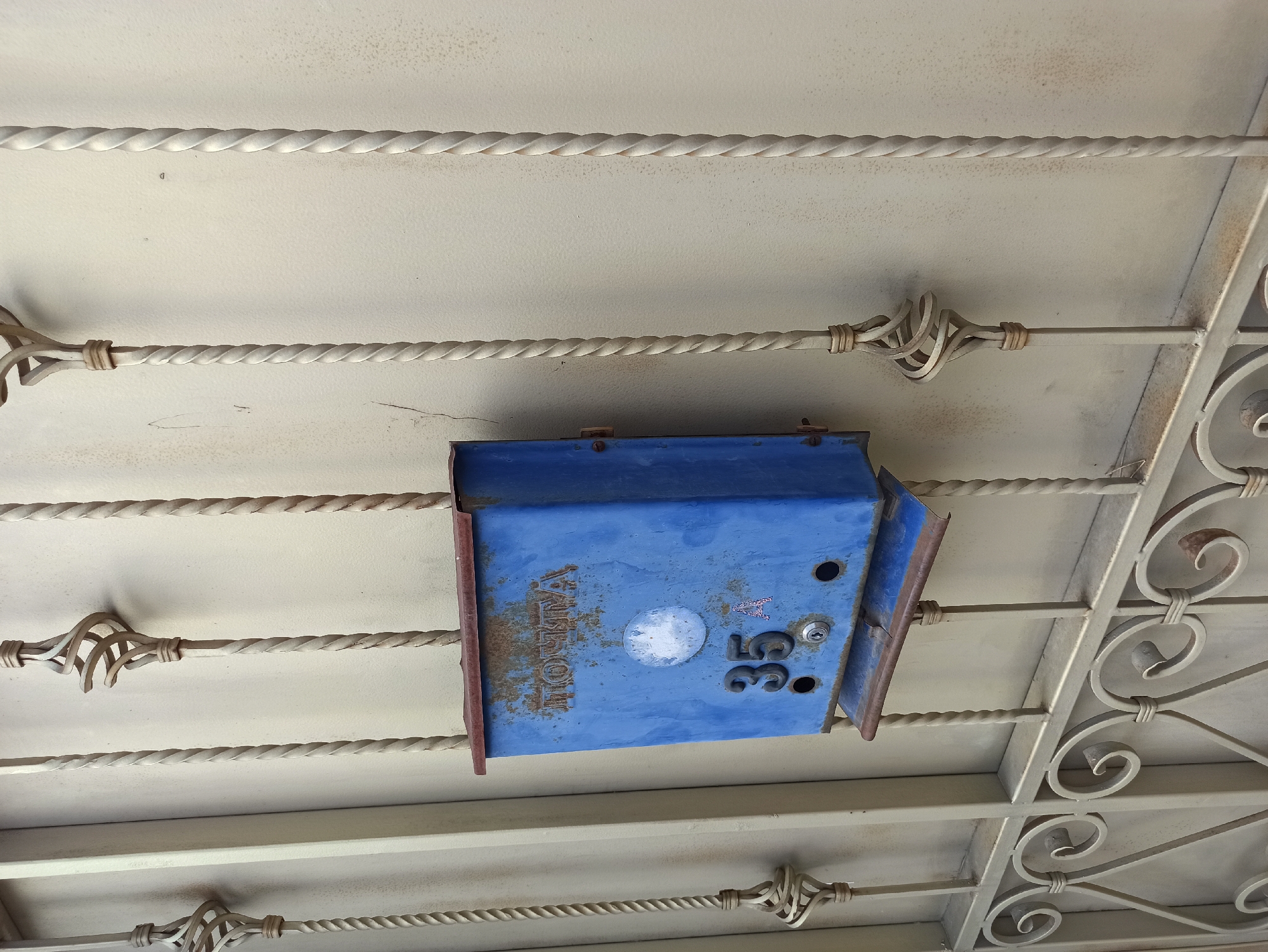 A blue metal mailbox is mounted on a light-colored wall behind a decorative metal fence. The mailbox is attached to the wall with visible screws. The word "ПОЧТА" (Post) is embossed in distressed, rusty letters on the front of the mailbox. Below the word, there is a circular white sticker or label, partially worn away. The number "35" is prominently displayed below the sticker, and a smaller "A" is visible to the right of it. Below the numbers, there are two circular openings, one above a keyhole, for mail delivery. The mailbox itself shows signs of rust and wear. The fence consists of vertical twisted metal bars and decorative scrolled elements at the bottom. The wall behind the mailbox appears to be textured and lightly stained. The scene is likely outdoors, given the presence of the fence and the overall appearance of the mailbox.