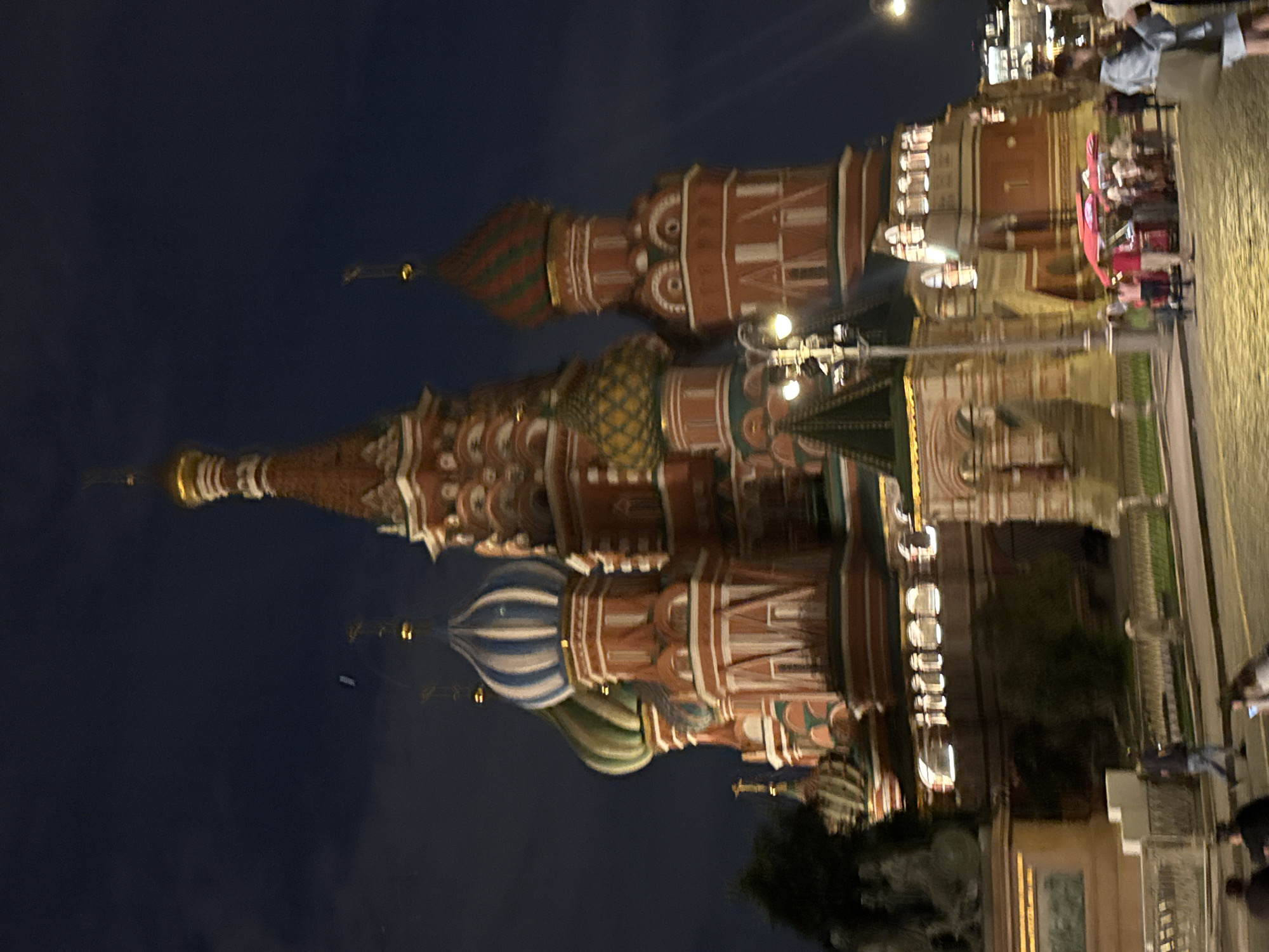 The image captures a nighttime view of Saint Basil's Cathedral in Moscow, Russia. The iconic onion domes of the cathedral are illuminated, set against a dark sky. In the foreground, several people are visible walking and standing on a cobblestone street. Some are gathered near a red-colored food stall. A lamppost with a light on stands prominently in the middle ground. The scene appears to be a public space with a wide pathway or road.