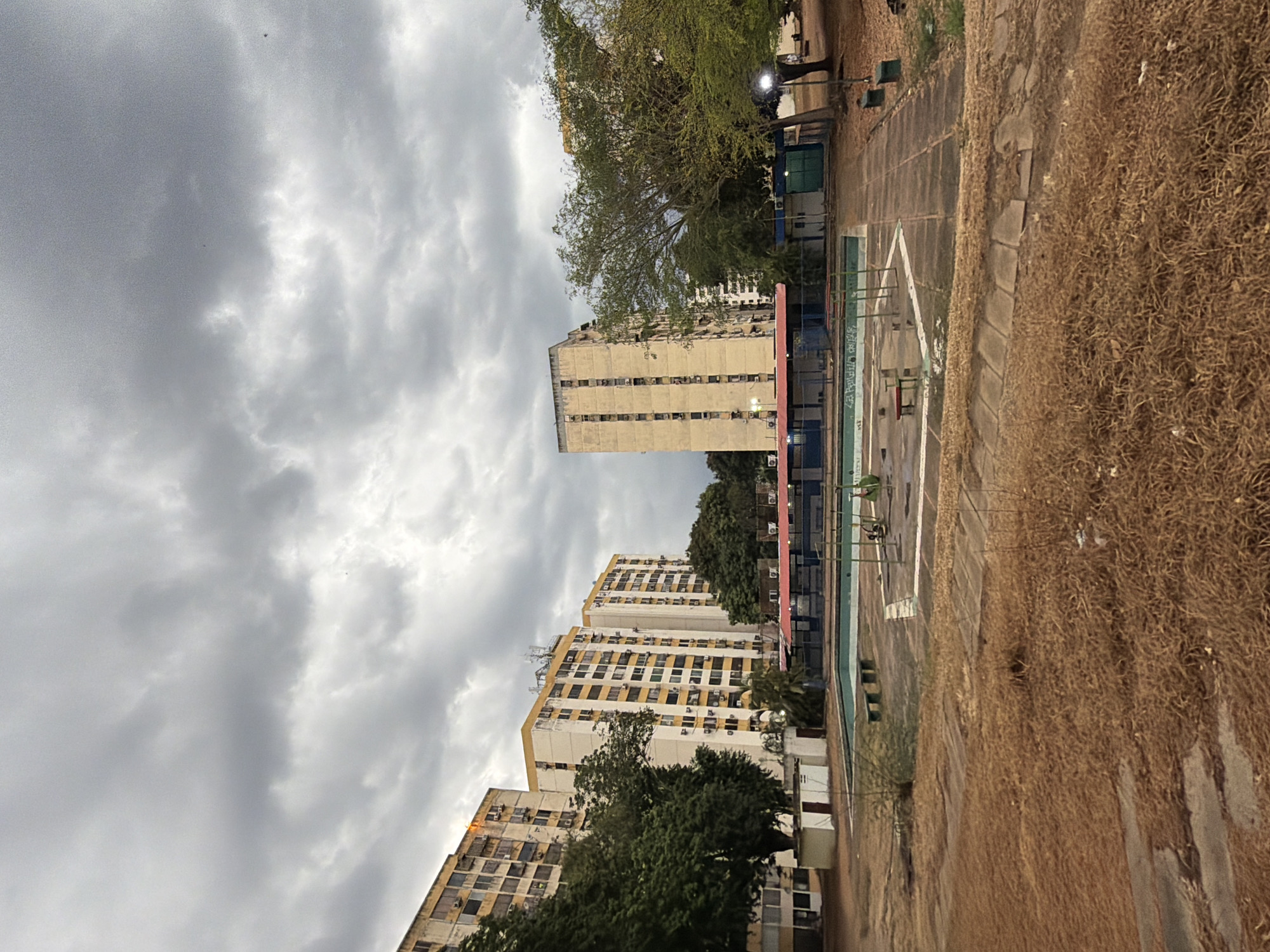 The image shows a cityscape in Ciudad Guayana, Venezuela. Several high-rise apartment buildings stand in the background under a cloudy sky. A sports court or recreational area lies in the foreground, with visible court markings and metal fencing. Overgrown vegetation surrounds the court area. The buildings appear to be residential, possibly part of a housing complex within the city.