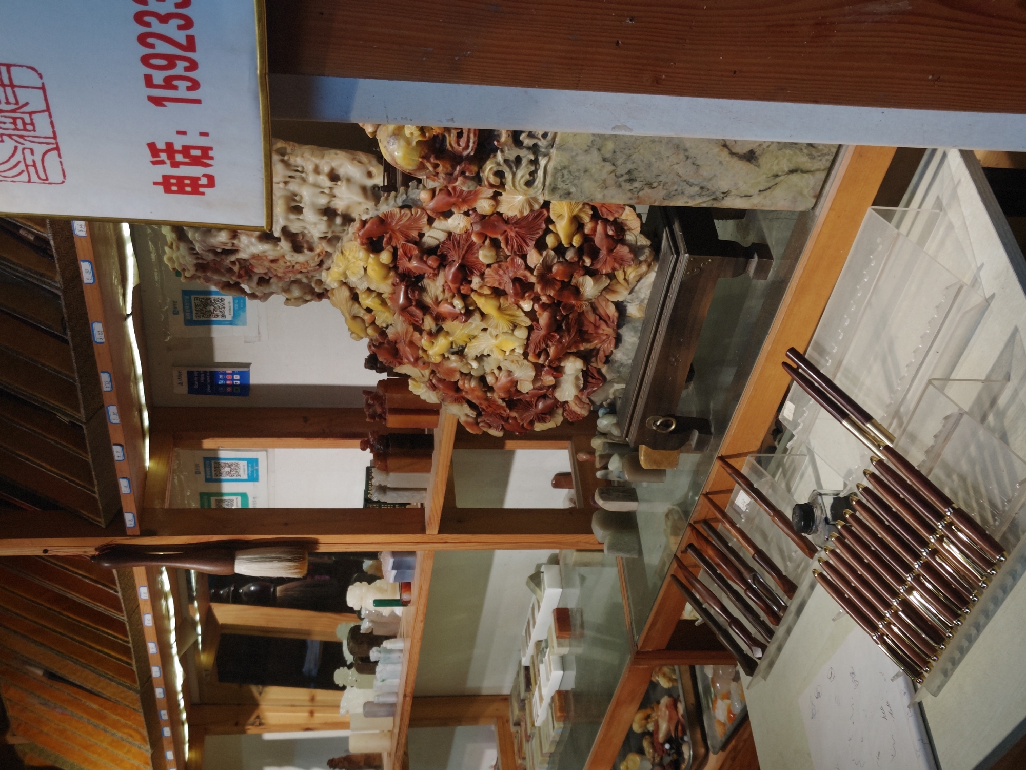 The image shows a shop display in Shapingba District, China. A glass-topped display case contains pens and small carved items. Shelves behind the display case contain various stone carvings and brushes. A sign displays Chinese characters and the phone number 159233.