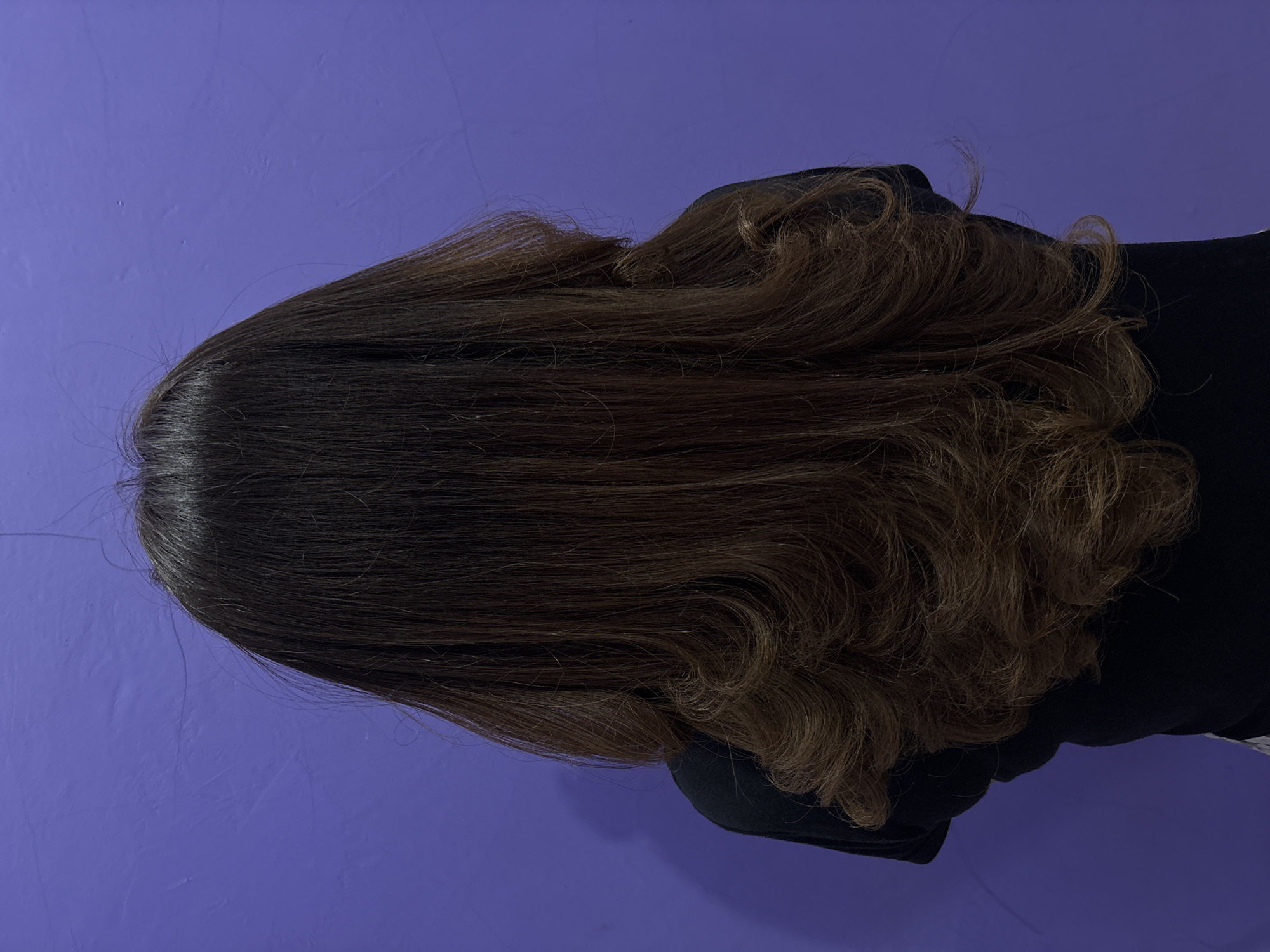 A person's back is shown, with their long, dark brown hair styled in loose waves. They are wearing a black t-shirt. The background is a solid purple wall. The image appears to be taken indoors, in a residential setting, possibly in North Jakarta, Indonesia.