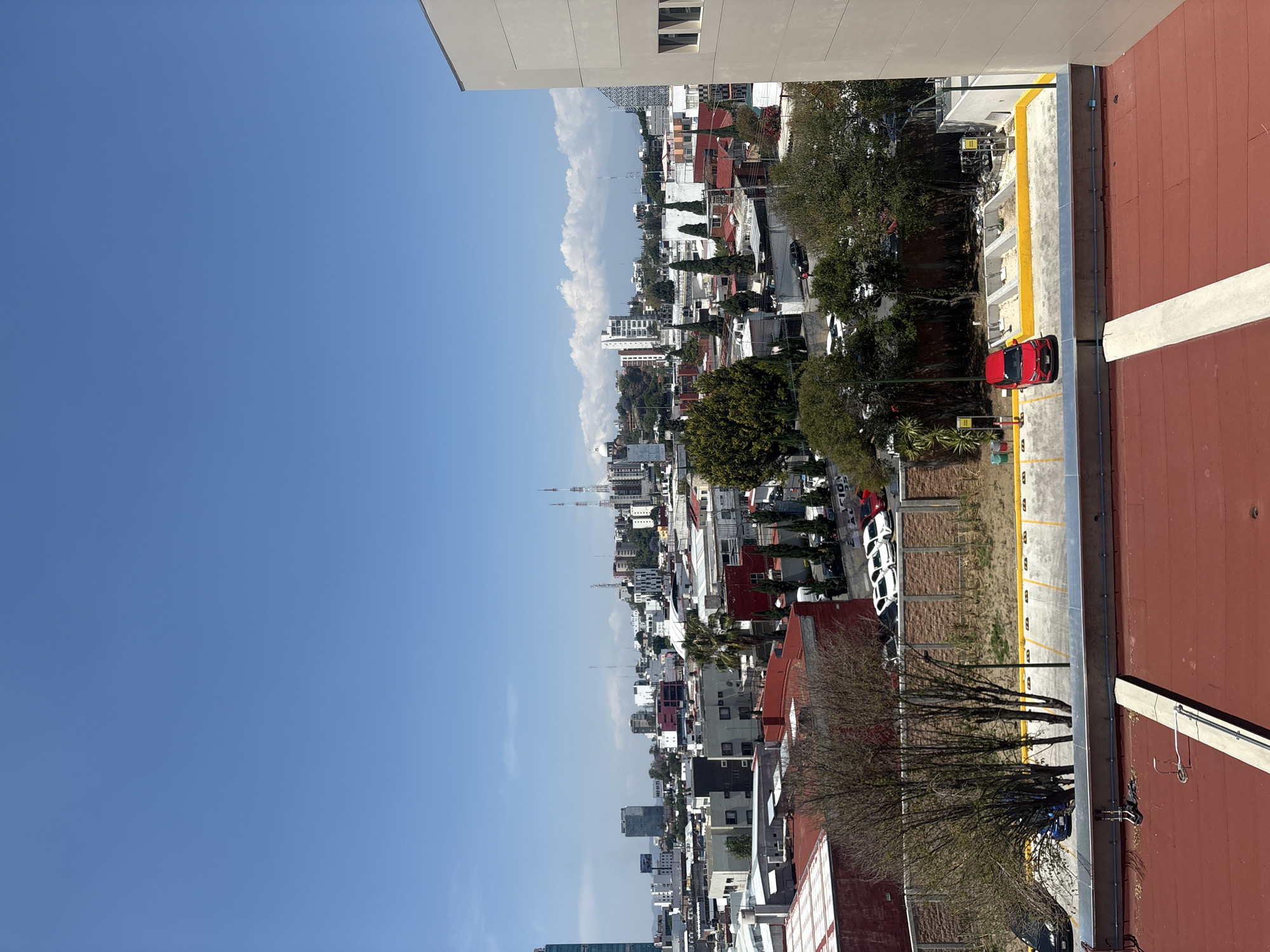 The image shows a cityscape in Puebla, Mexico, taken from an elevated perspective. The sky is a clear blue with some clouds in the distance. The cityscape consists of a mix of residential and commercial buildings, interspersed with trees and greenery. Buildings exhibit a variety of architectural styles and colors, with many having red-tiled roofs. There are multiple vehicles parked along the street. Visible in the background are tall antenna towers. The foreground includes part of the building from which the photo was taken, featuring a flat red-tiled surface and a painted white line.