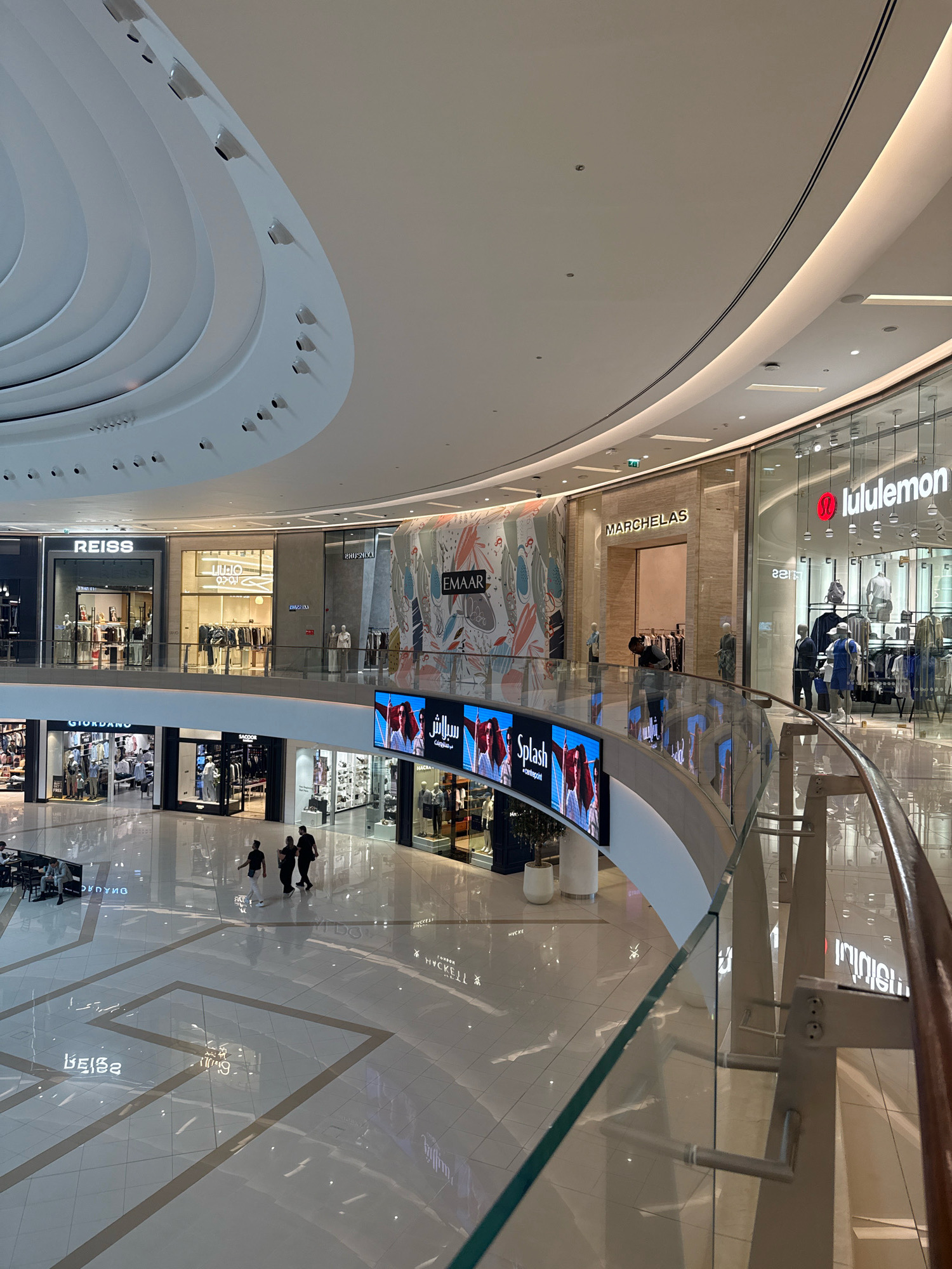 The image shows a wide shot of the interior of a large, modern shopping mall in Dubai, United Arab Emirates. The mall features multiple levels, a high, curved ceiling with integrated lighting, and glass railings overlooking the ground floor. Several shops are visible on the upper and lower levels, including "REISS," "GIORDANO," "MARCHELAS," and "Lululemon." Digital screens displaying advertisements are mounted on a curved structure in the foreground. People are seen walking on the ground floor and on the upper level. The mall's floor is reflective, showing the lights and store facades.