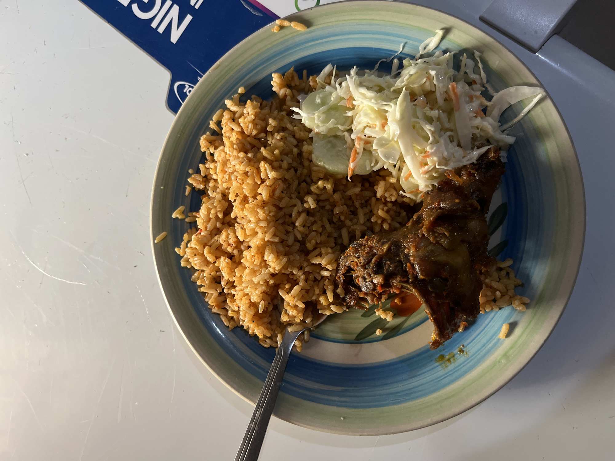 A close-up overhead shot shows a plate of jollof rice with chicken and coleslaw. The jollof rice is a reddish-orange color and fills most of the plate. To the left of the rice is a piece of dark brown, seasoned chicken. A small portion of coleslaw, made of shredded cabbage and carrots with a creamy dressing, sits to the right of the rice. A fork is visible in the upper left corner, resting on the rice. The plate has a decorative rim with blue, green, and brown stripes. In the background, a blue object with white lettering that reads "NIGERIA" is partially visible. The scene is set on a white, slightly textured surface that appears to be a table. The lighting suggests an indoor setting, possibly a restaurant or home. The image does not provide any specific information about the city of Bukuru, Nigeria.