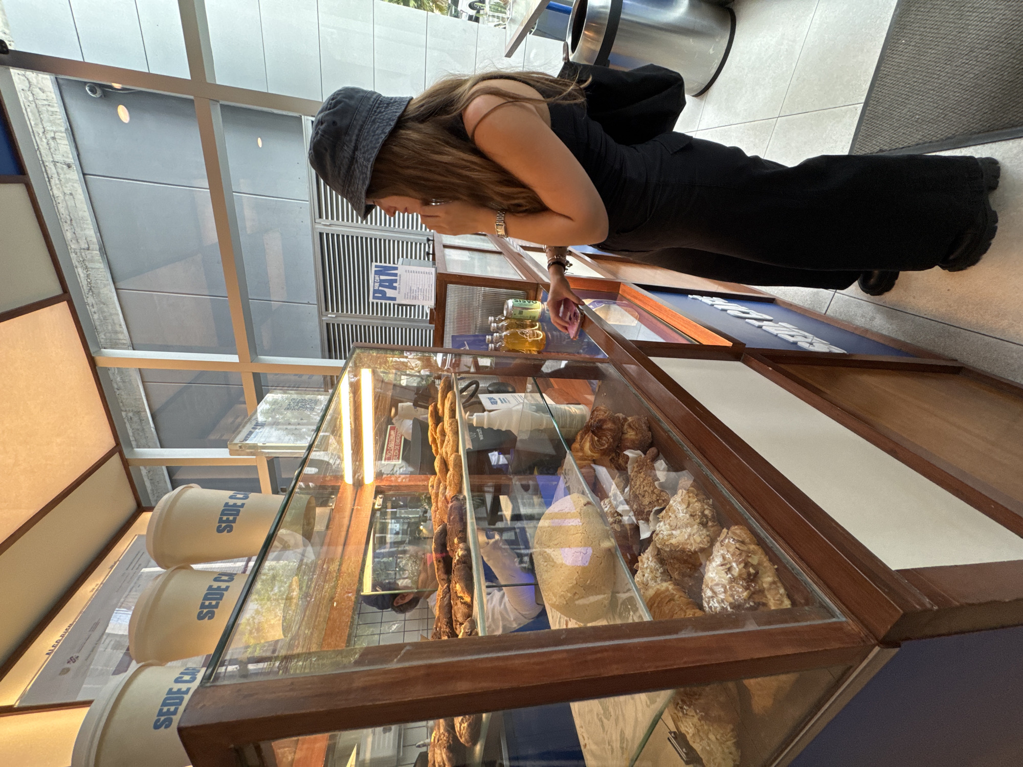 Here's a description of the image:
A young woman wearing a dark bucket hat and a black dress stands in front of a bakery display case. She is looking down at the pastries and items inside the case. The bakery case is made of wood and glass, and it contains various types of pastries and bread. There are also some cups with the words "SEDE CA" visible through the glass.
In the background, there's a glimpse of the building's interior and a trash can. The lighting is bright, suggesting it's daytime. There is a sign hanging that reads "PAN"
