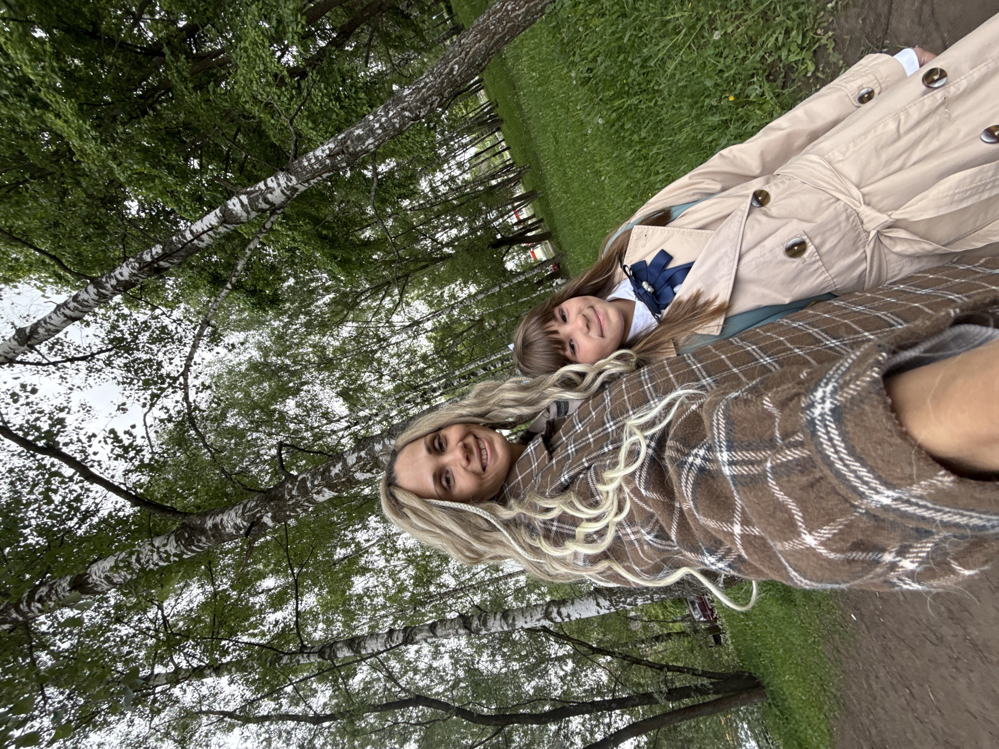 The image depicts two females lying on the ground in Rusanovo, Russia, amidst a birch tree forest. The younger female is wearing a white shirt, a blue bow, and a tan trench coat. The older female is wearing a brown plaid shirt and has long blonde hair with braids. They are lying on the ground, which is covered in grass and dirt. The background consists of many birch trees with green leaves. The sky is overcast.