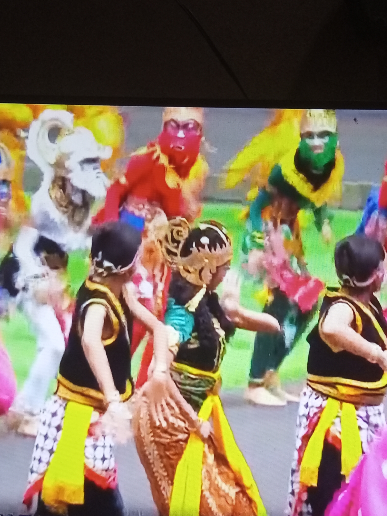 The image captures a lively street performance in Banyudono, Indonesia, featuring several individuals adorned in vibrant, traditional costumes. The central figures are dressed in black vests with gold trim, embellished with yellow sashes. Some wear elaborate headdresses, including one resembling a white creature and another in gold with intricate patterns. The performers are engaged in a dynamic dance, with their arms outstretched and movements suggesting a celebratory atmosphere. The background reveals more performers in similarly colorful attire, with some wearing masks of red and green, and others in bright yellow and blue outfits. The ground is paved, indicating an outdoor setting, possibly a public square or street in the town of Banyudono. The overall scene is one of cultural festivity and energetic dance.