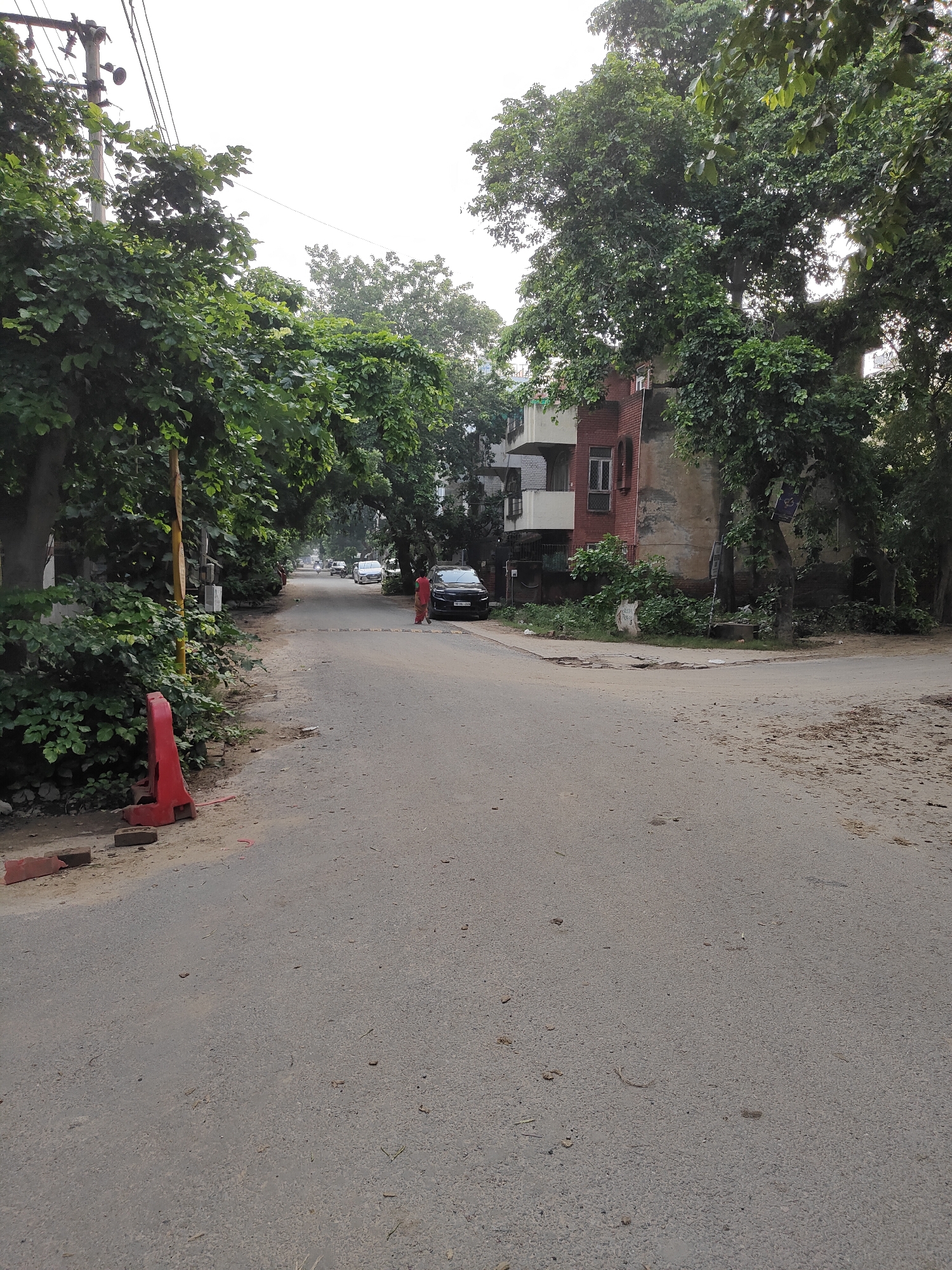 The image captures a residential street in Gurgaon, India, on a cloudy day, likely in the late morning or early afternoon. The road, paved with asphalt, curves gently into the distance, flanked by lush green trees and foliage. A few vehicles are visible on the road, including a black SUV parked on the right side and a white van further down. A woman in a red kurta is walking on the right side of the road.
On the left side of the street, a red construction barrier and some bricks are placed near the roadside, indicating ongoing work or maintenance. A yellow pole, possibly a utility marker or part of a traffic sign, stands beside the barrier. Behind the barrier, dense green bushes and trees create a natural boundary.
To the right, a multi-story building with a red brick facade and white balconies is partially visible, partially obscured by the dense canopy of trees. The building appears to be residential. The presence of mature trees lining the street suggests an established neighborhood. The sky is overcast, creating a diffused light that softens the scene. The overall atmosphere is calm and typical of a quiet suburban street. No discernible text is visible in the image.