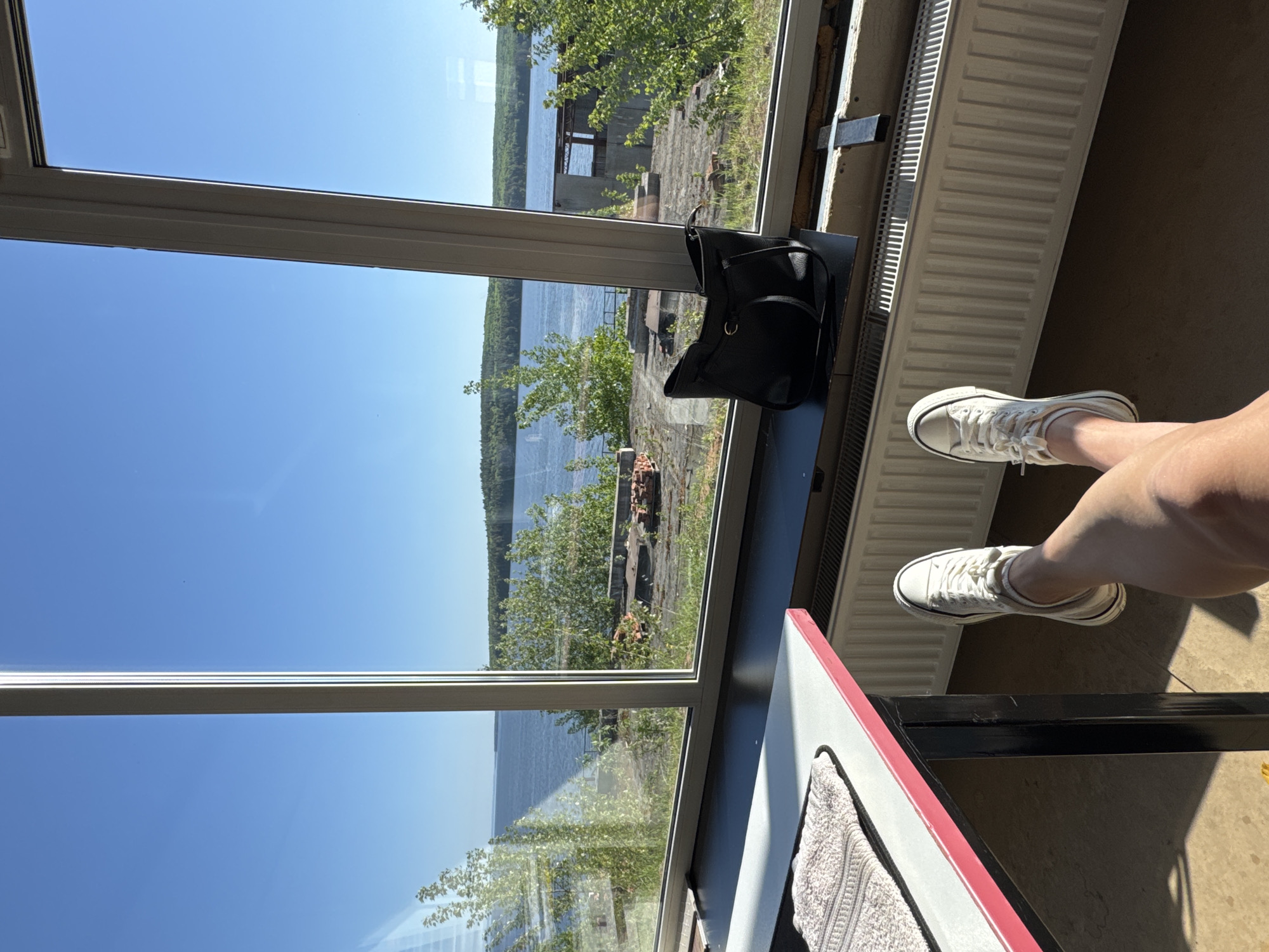 The image depicts an indoor scene with a view of a body of water, potentially a lake or river. The sky is clear and blue.
In the foreground, a black purse rests on a window sill. Adjacent to the window, part of a white radiator is visible. A person's legs and feet, wearing white sneakers, are positioned to the right of the radiator. A table with a red edge and a towel on it is also in the shot.
Based on the scenery, it is plausible that the location could be Chaikovsky, Russia, due to its proximity to the Kama River and associated reservoirs, which create numerous waterside environments.