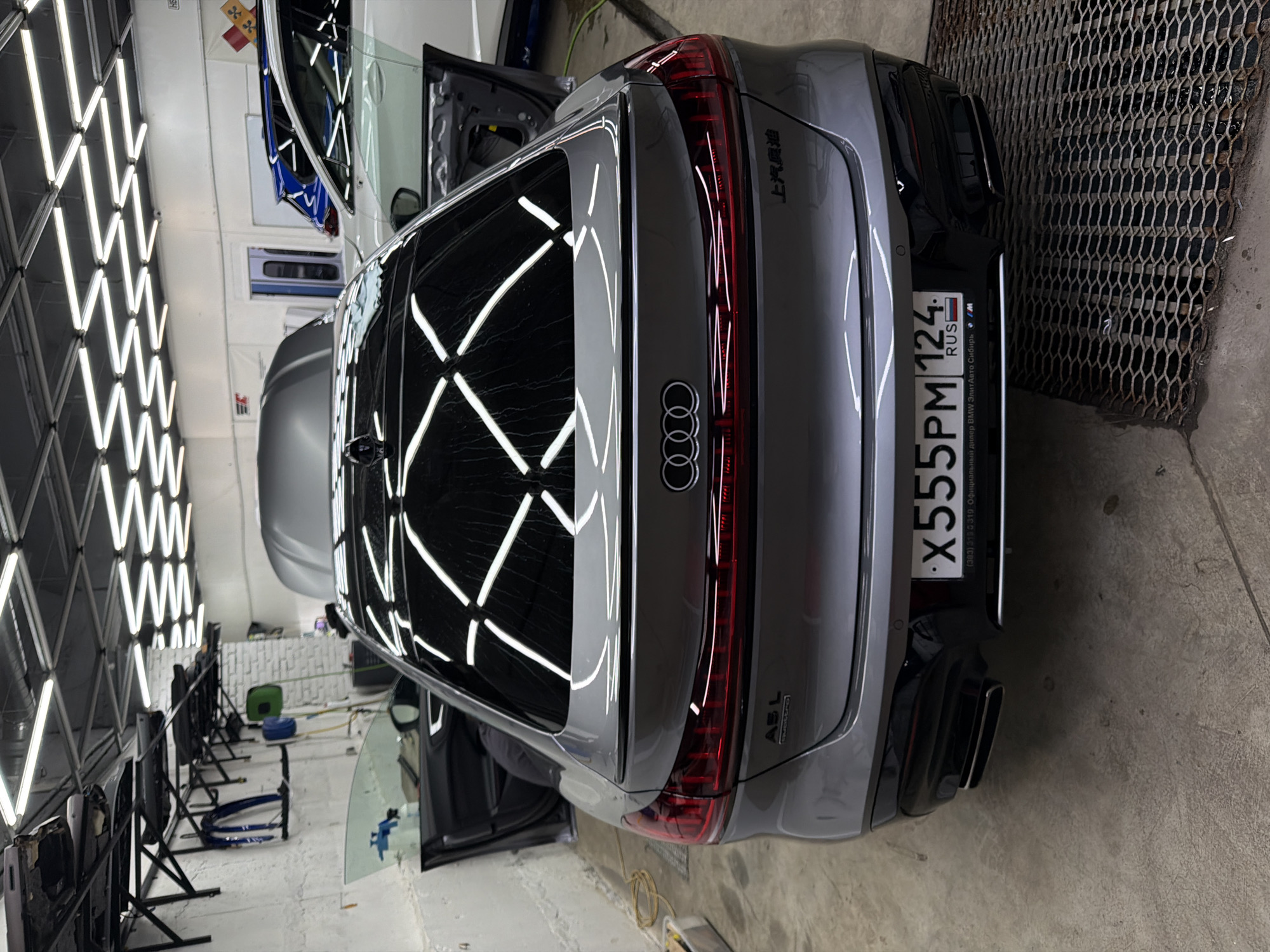 The image shows the rear of a gray Audi station wagon-style car, likely in a detailing or car customization shop. The car's license plate is "X555PM 124," indicating registration in the Krasnoyarsk region of Russia. A dealership sticker from "Элит Авто Сибирь," an official BMW dealer, is also present on the license plate frame. The car is inside a well-lit workshop, with a rack of lights reflecting off the rear window. The rear of another vehicle (likely a white sedan) is visible behind the Audi. Several car doors are detached from vehicles and are on stands, suggesting the shop focuses on repairs or customization. A perforated metal grate is visible on the right side of the frame, indicating possible drainage and the presence of water. The letters “ASL”, quattro, and the Chinese lettering “上汽奥迪” are visible on the back of the car.