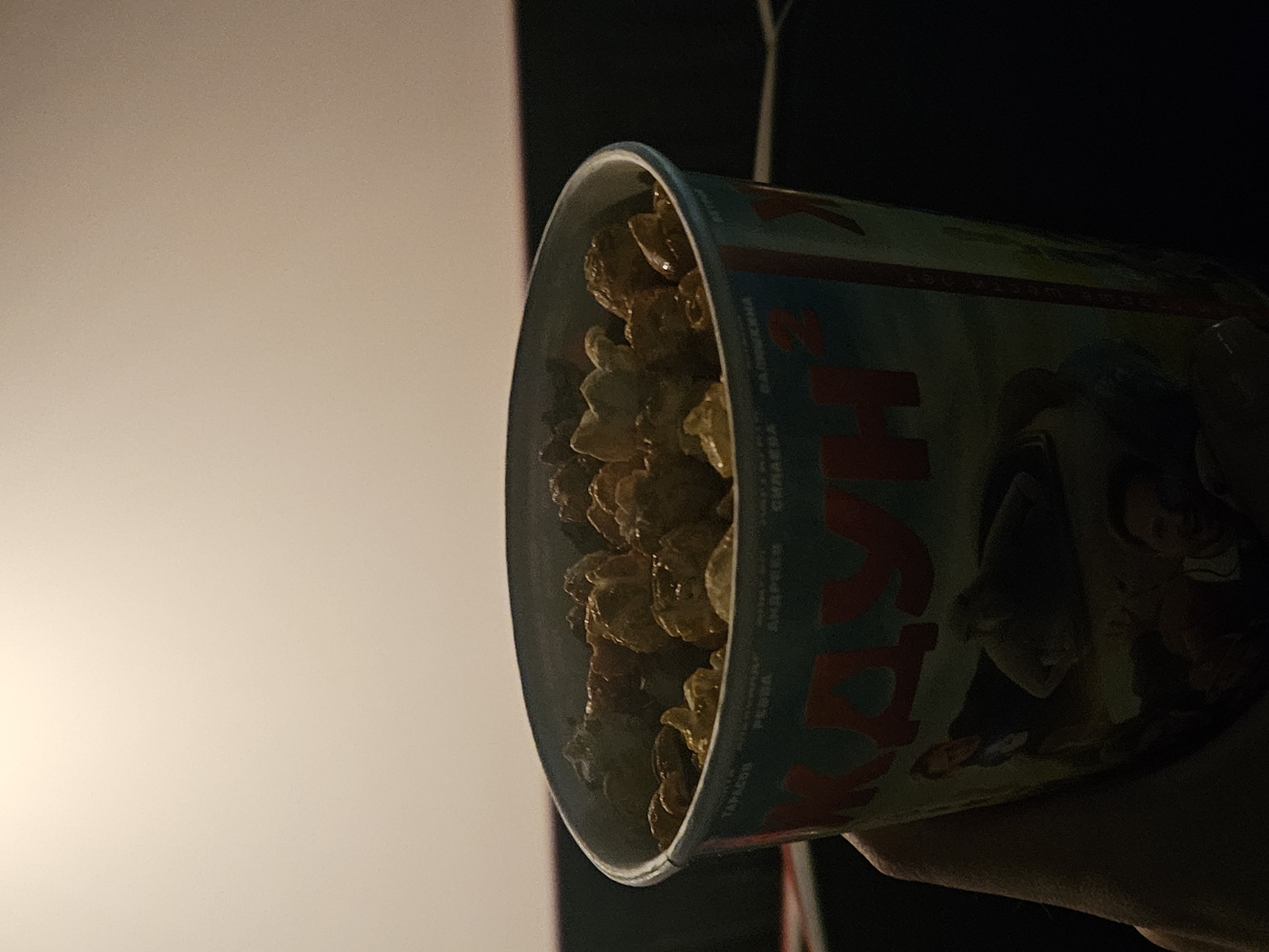 Here is a forensic description of the image:
The image depicts a container of popcorn held in what appears to be a person's hand, likely in a movie theater. The popcorn is caramel-colored, and the container is decorated with images and text in what looks like Russian. The visible word on the container is "ШАХУН", which is Russian for Shakhun. The location is likely a movie theater in Shakhunya, Russia. The background shows a dimly lit area with a partially visible projection screen, further indicating the movie theater setting.