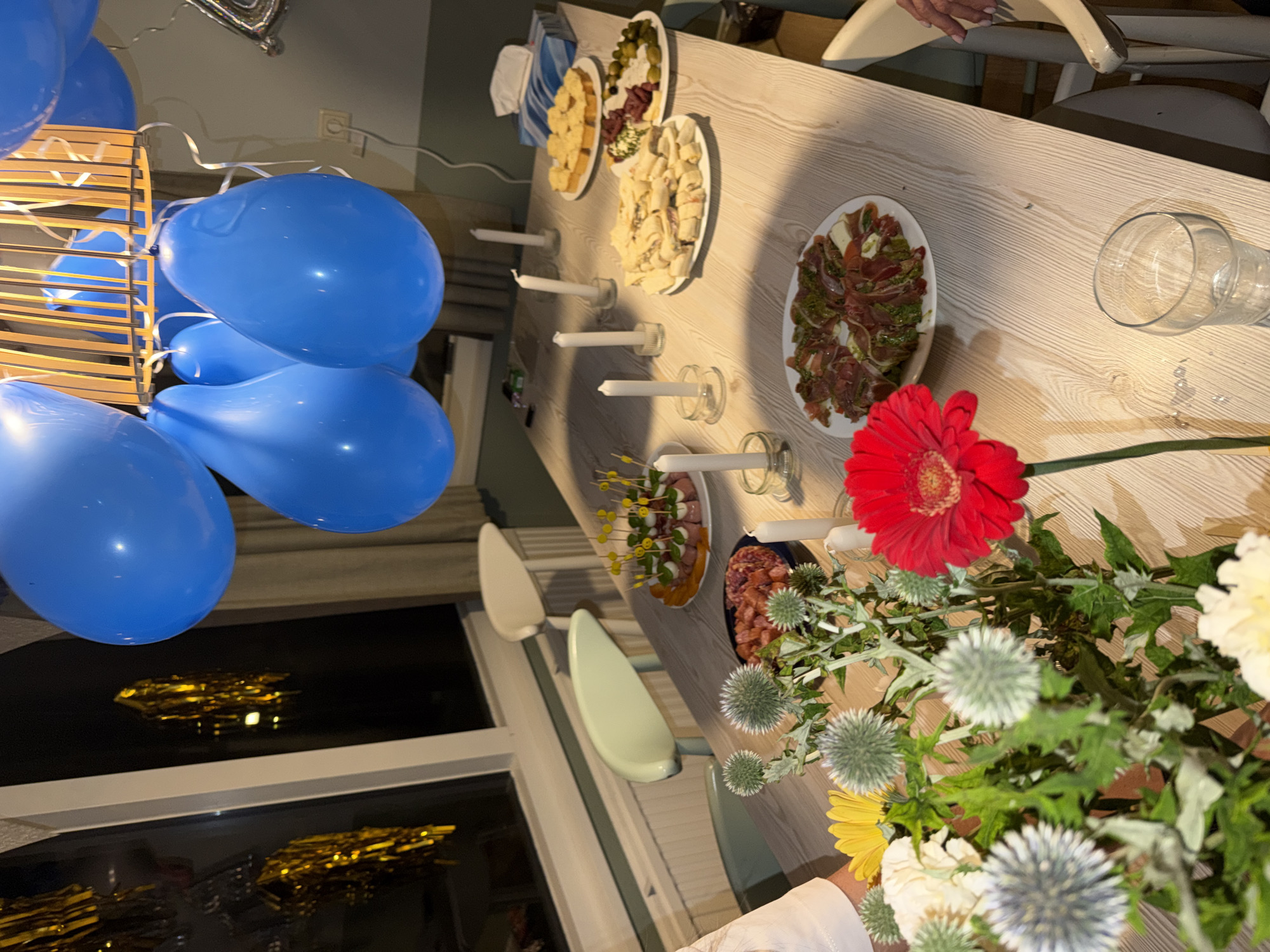 This image depicts a table set for a celebration, likely a birthday party, in Heijen, Netherlands. A wooden table is laden with various food items, including sandwiches, small bites on skewers, and sliced meats. Several white candles in glass holders and a bouquet of flowers, featuring a prominent red gerbera and thistles, add to the festive atmosphere. Blue balloons are prominently displayed, hanging from what appears to be a ceiling fixture. The room is dimly lit, with reflections visible in a window in the background. The presence of chairs suggests guests are or will be present. No individuals are clearly visible in the frame, however, a hand is partially visible on the right side of the image.
