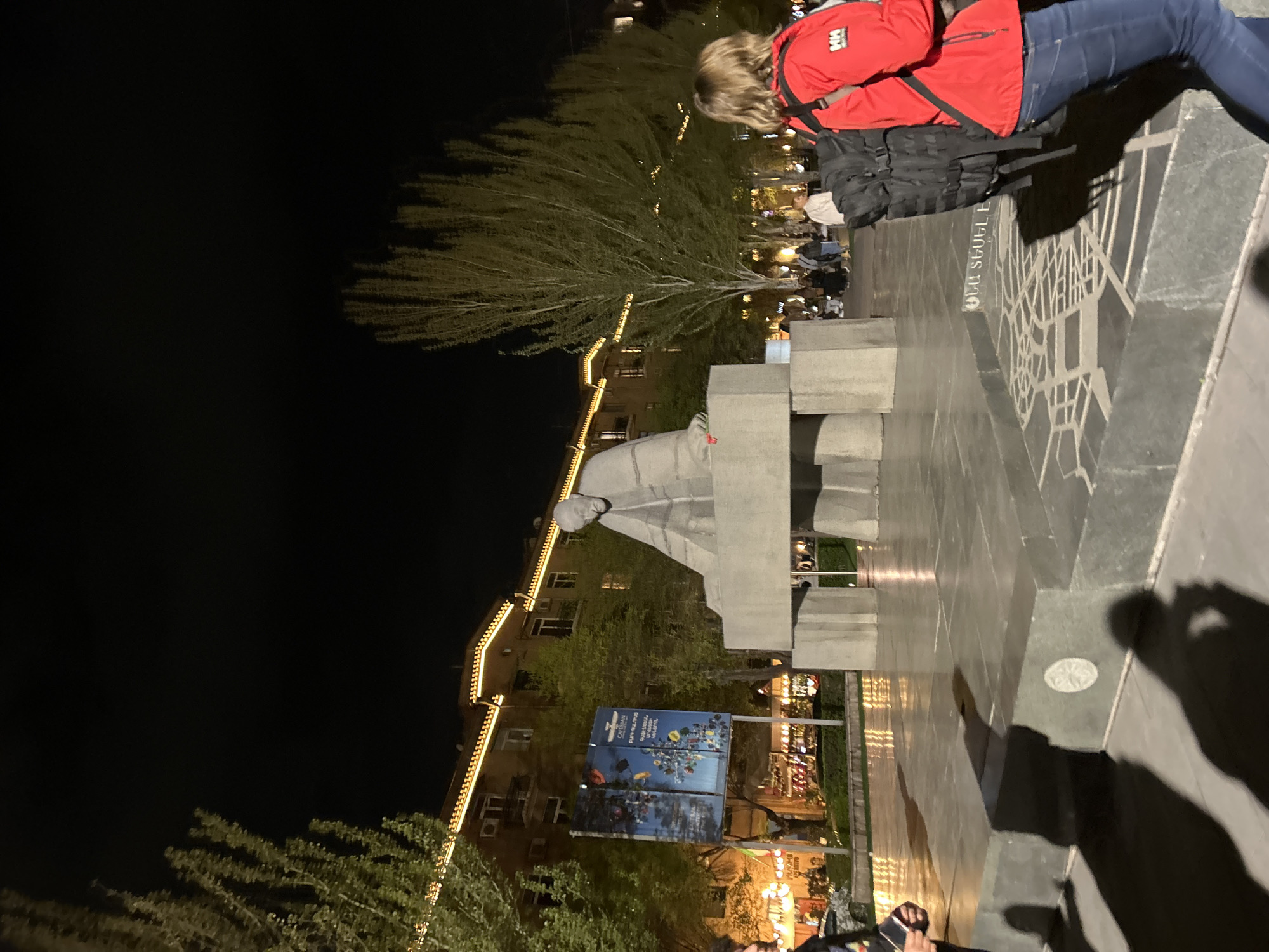 Here is a detailed description of the image:
The image captures a nighttime scene at the Matenadaran Mesrop Mashtots Institute of Ancient Manuscripts in Yerevan, Armenia. The monument of Mesrop Mashtots is prominently featured. The stone surfaces around the monument appear to have a map engraving. A person in a red jacket and blue jeans is either sitting on the edge of the monument area. Additional people are walking around the monument. Buildings decorated with lights are present, and a sign with blue panels displaying images or information is visible. The scene is well-lit, highlighting the architecture and the monument.