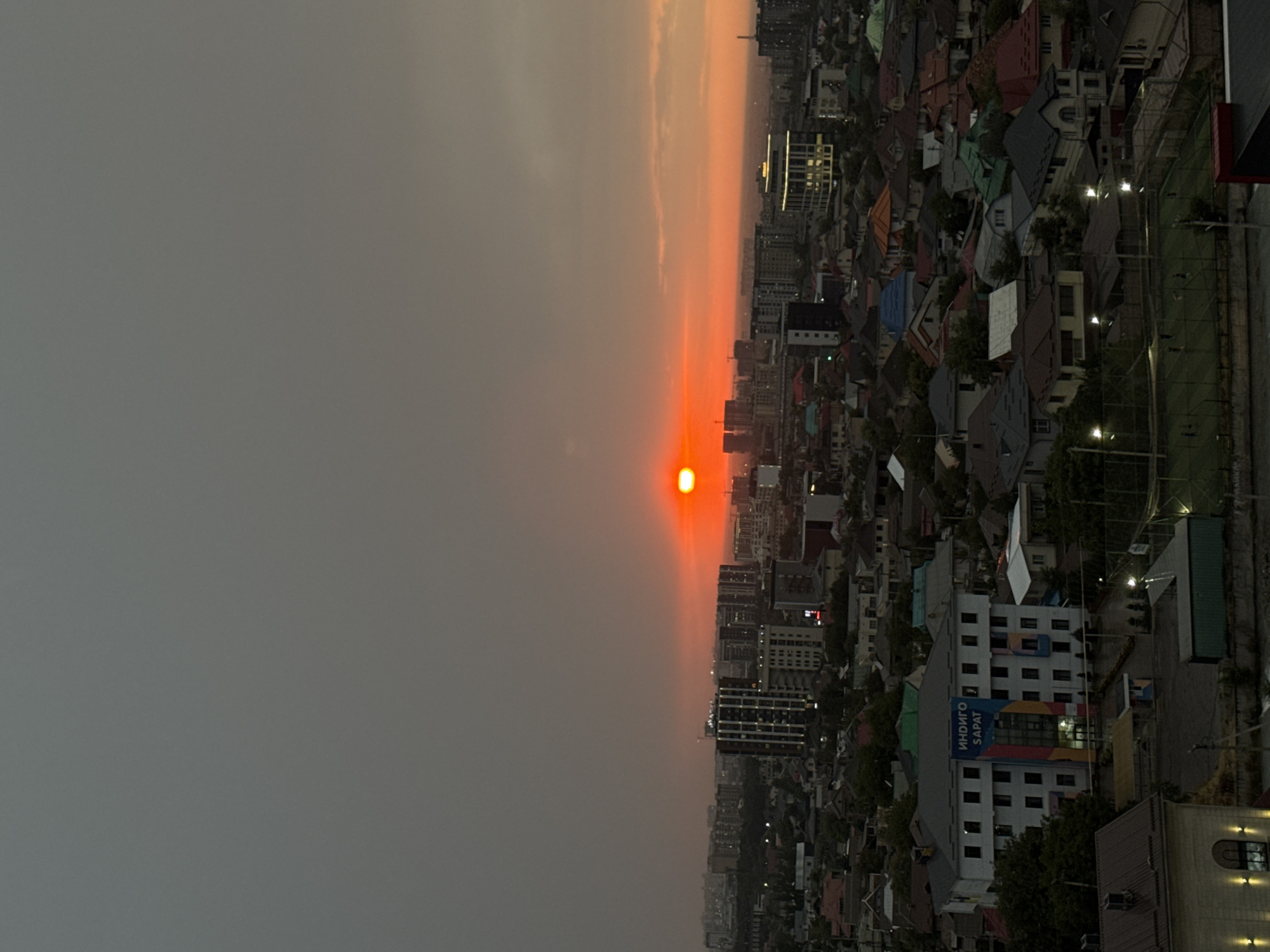A vertical, medium-shot photograph captures a hazy sunset over a residential and commercial area in Bishkek City, Kyrgyzstan. The sky is a dull gray with a bright, orange sun peeking through the haze on the right side of the frame, casting an orange glow on the clouds and horizon. Below the sky, a dense urban landscape unfolds, characterized by a mix of low-rise residential buildings with pitched roofs in various colors, and taller, modern commercial buildings.
One prominent building in the foreground, to the left of center, is a white, multi-story structure with numerous windows and a banner hanging vertically on its facade. The banner displays blue text, appearing to read "ИНДИГО SAPAT". Another multi-story building, to the right of the banner building, is illuminated from within. In the distance, more city structures are visible, some appearing as silhouettes against the glowing horizon.
A sports field, possibly a soccer field, with players can be seen in the lower right quadrant of the image. The overall impression is of a bustling city during twilight, with the hazy sunset creating a somewhat muted and diffused atmosphere. The image appears to have been taken from an elevated vantage point.