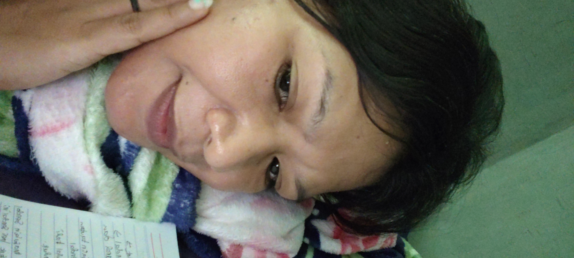 The image is a close-up, low-angle, full-body shot of a young person lying down, with their head to the right. They have dark hair, dark eyes, and light-colored skin. The person is wearing a ring on their left index finger. They are resting on a colorful blanket with stripes of white, pink, blue, and green. To the left of the person, a corner of a notebook with handwritten notes is visible. The background is a plain green wall. The scene appears to be indoors. There are no discernible landmarks or elements that would place the image in Sika Makmur, Indonesia.