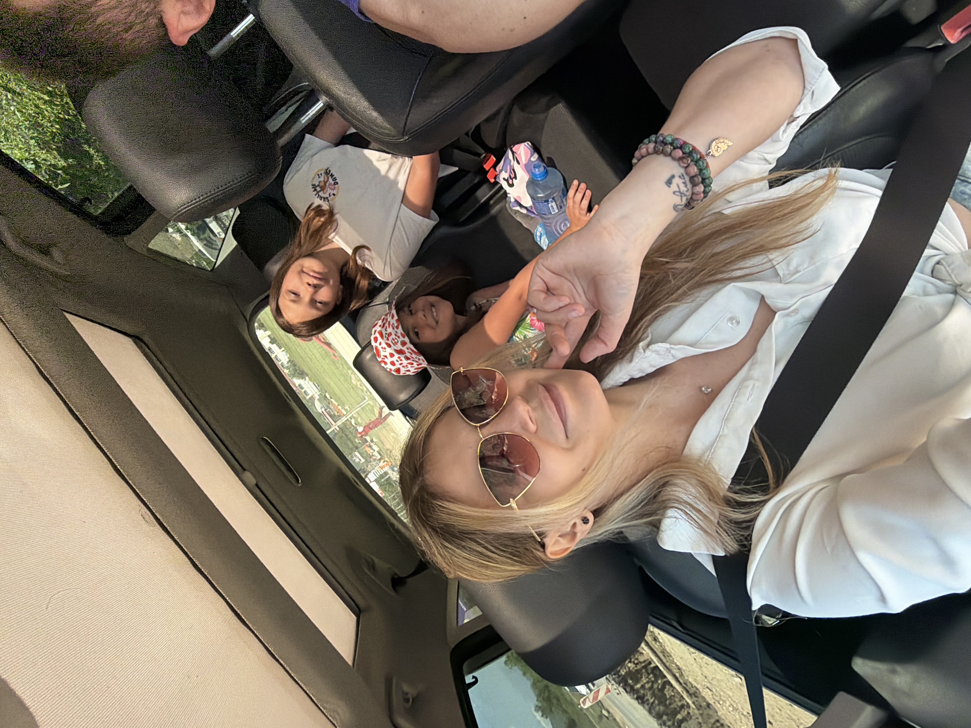 The image shows a woman in the front passenger seat of a vehicle, wearing sunglasses and a white shirt, with a seatbelt across her chest. In the back seat, there are two children. One child is wearing a white shirt with a graphic, and the other is wearing a floral patterned shirt and a red and white headband. A water bottle and a patterned bag are also visible in the back seat. The image was taken in Prijevor, Montenegro.