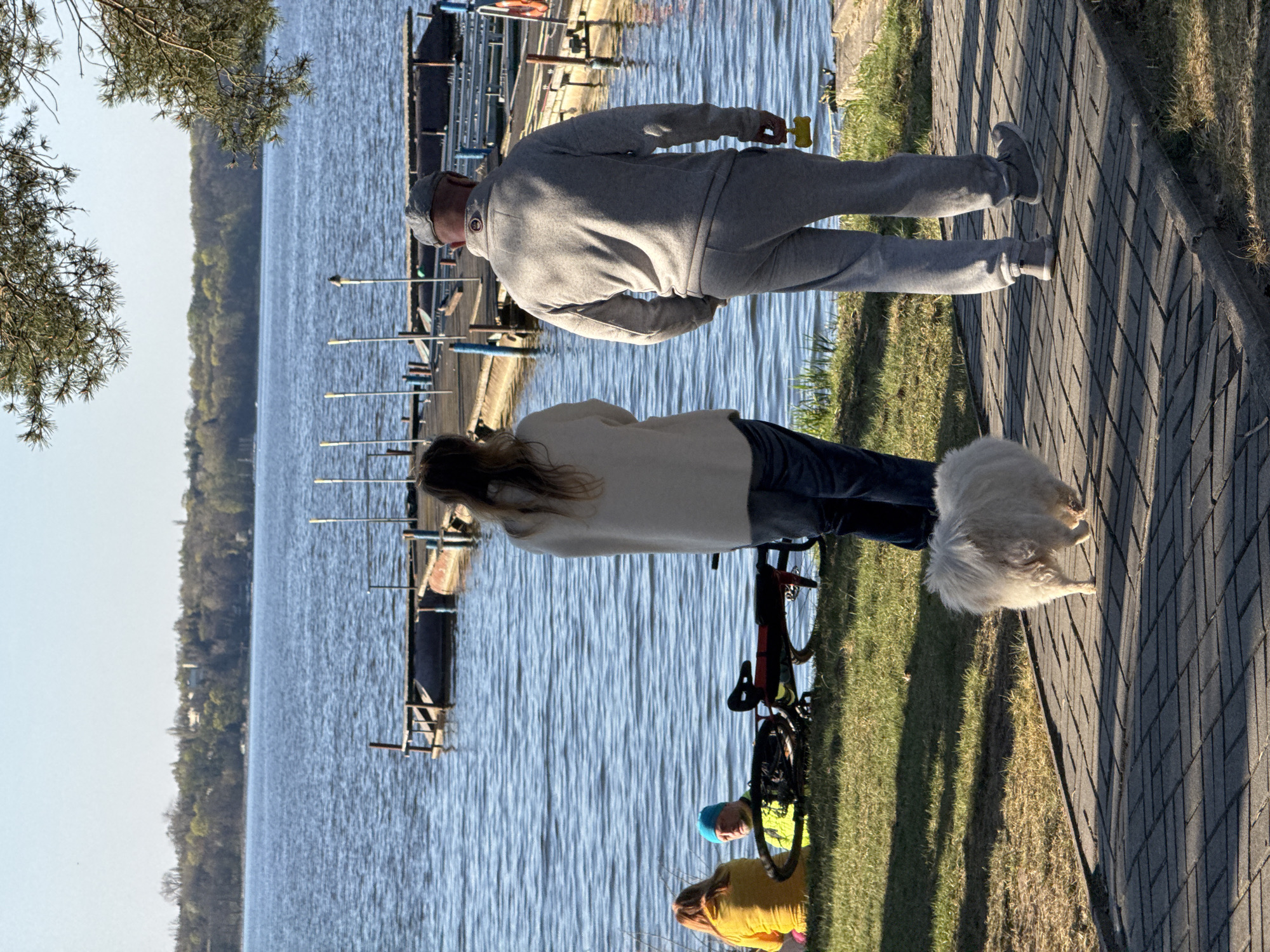The image depicts a scene by the Pilica River in Białobrzegi, Poland. An older man in a gray sweatsuit walks along a paved path with a yellow object in his hand. A woman with long hair, wearing a beige sweater and jeans, stands nearby with a bicycle. A fluffy white dog is next to her. Closer to the river, two children are present; one in a yellow shirt and another wearing a blue hat. The river is to the left of the frame, with docks and boat-related structures extending into the water. Trees line the opposite bank.