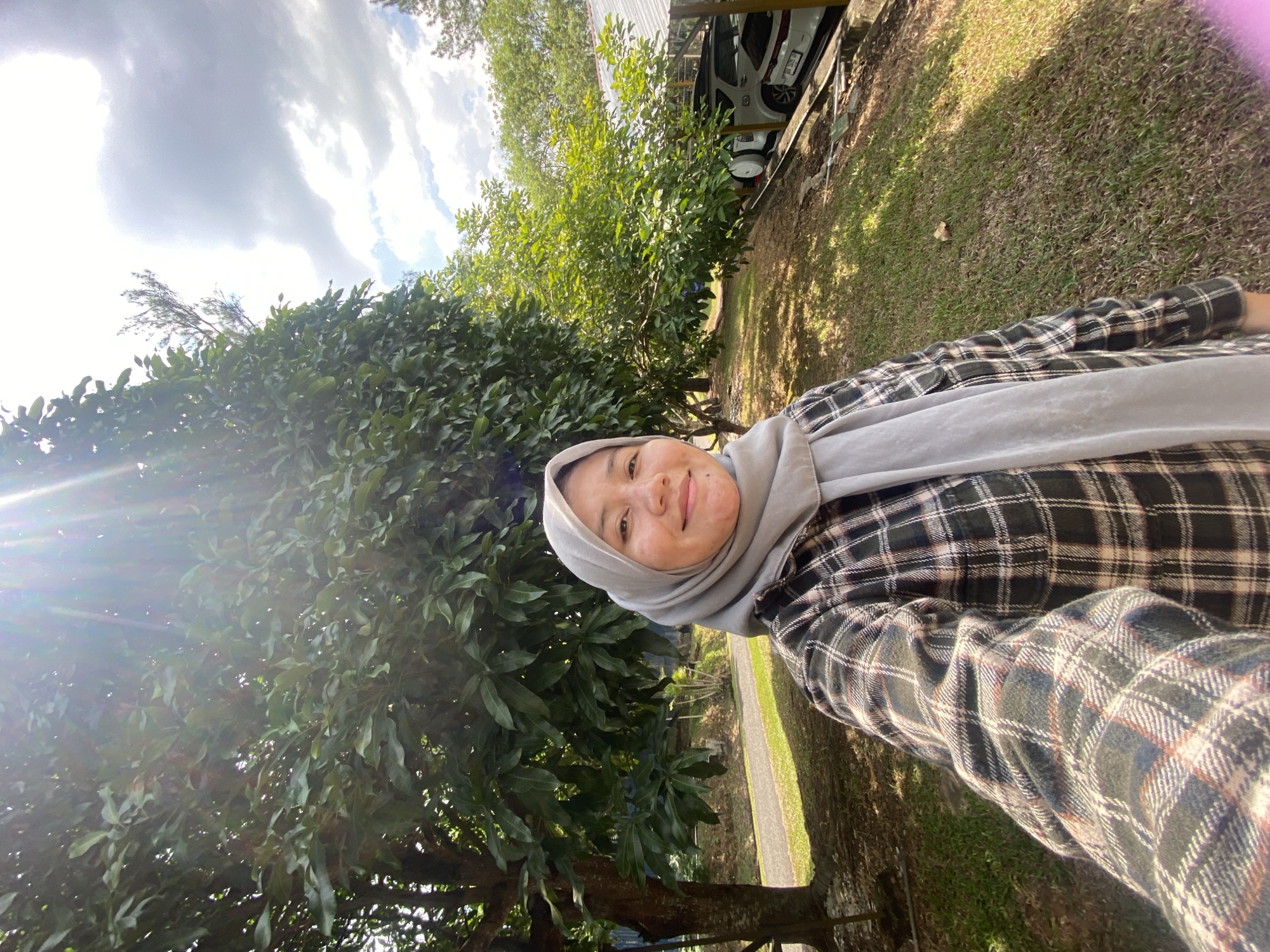 A selfie is taken by a person wearing a grey hijab and a plaid shirt, standing outdoors in what appears to be a park or recreational area. The person is smiling at the camera. In the background, there are several large trees with dense green foliage. Sunlight filters through the leaves, creating lens flare and highlighting parts of the scene. To the right of the frame, a covered parking structure with a white SUV and part of another vehicle is visible. The ground is covered with grass and some dry leaves. The sky above is partly cloudy with bright sunlight. The location is identified as Banjarbaru, Indonesia.