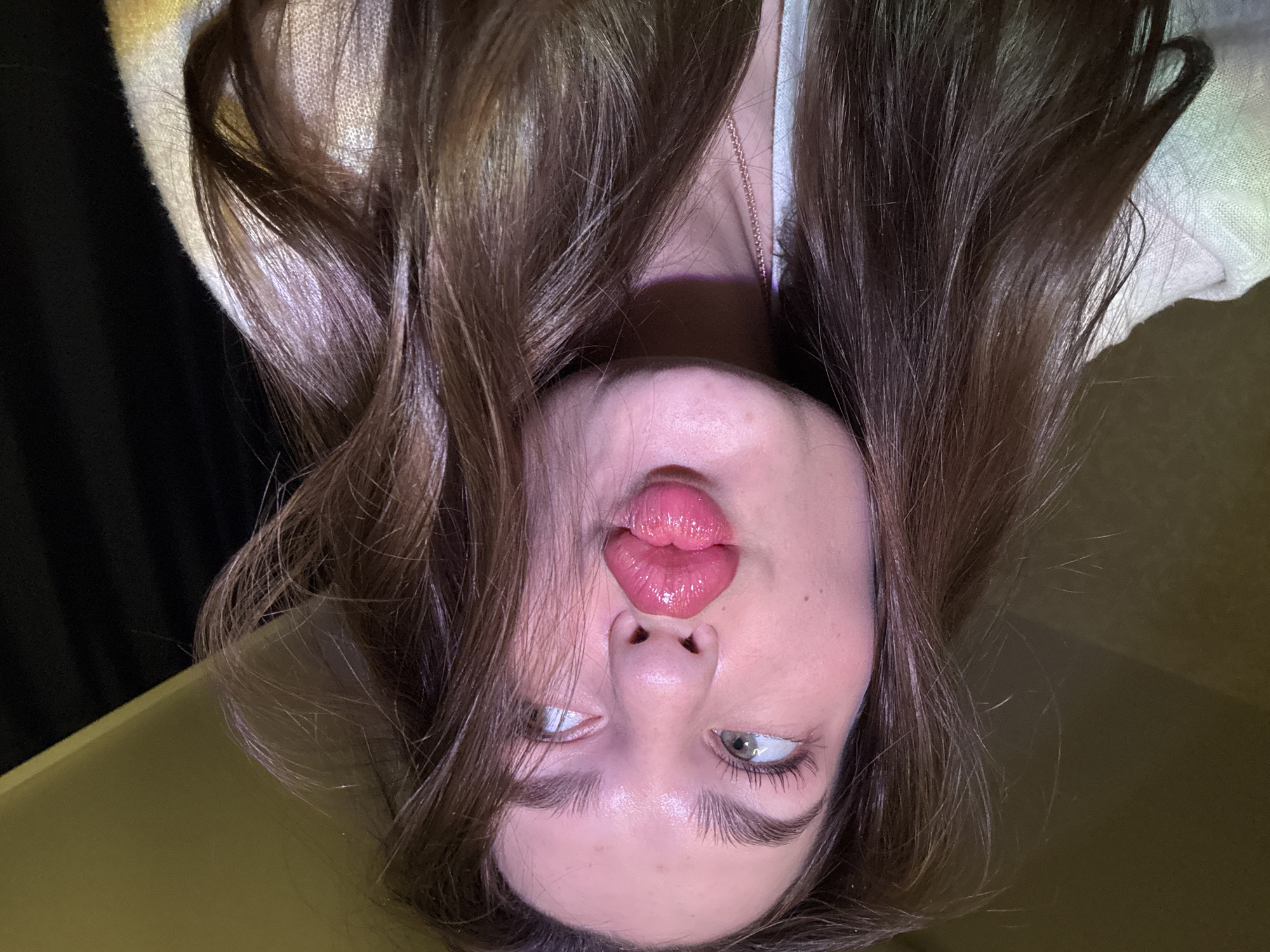 A person is upside down with long, dark hair draped around their face. They are making a kissing face with glossy lips. Their eyes are looking upwards. They appear to be indoors, with a dark background behind their hair at the top of the frame, and a light-colored surface visible at the bottom. There is no discernible evidence to place the location in Chaikovsky, Russia.