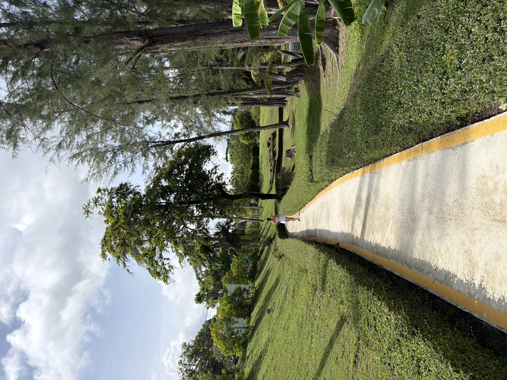 A sideways-oriented photograph depicts a person walking along a concrete path in a grassy, tree-lined area, possibly in Choeng Thale, Thailand. The path has a yellow border. To the right of the path is thick green shrubbery. To the left, there are trees of varying sizes and species, and a grassy slope that leads to more trees and some buildings further away. The sky is partly cloudy. The person on the path is wearing light-colored clothing and is walking away from the viewer.