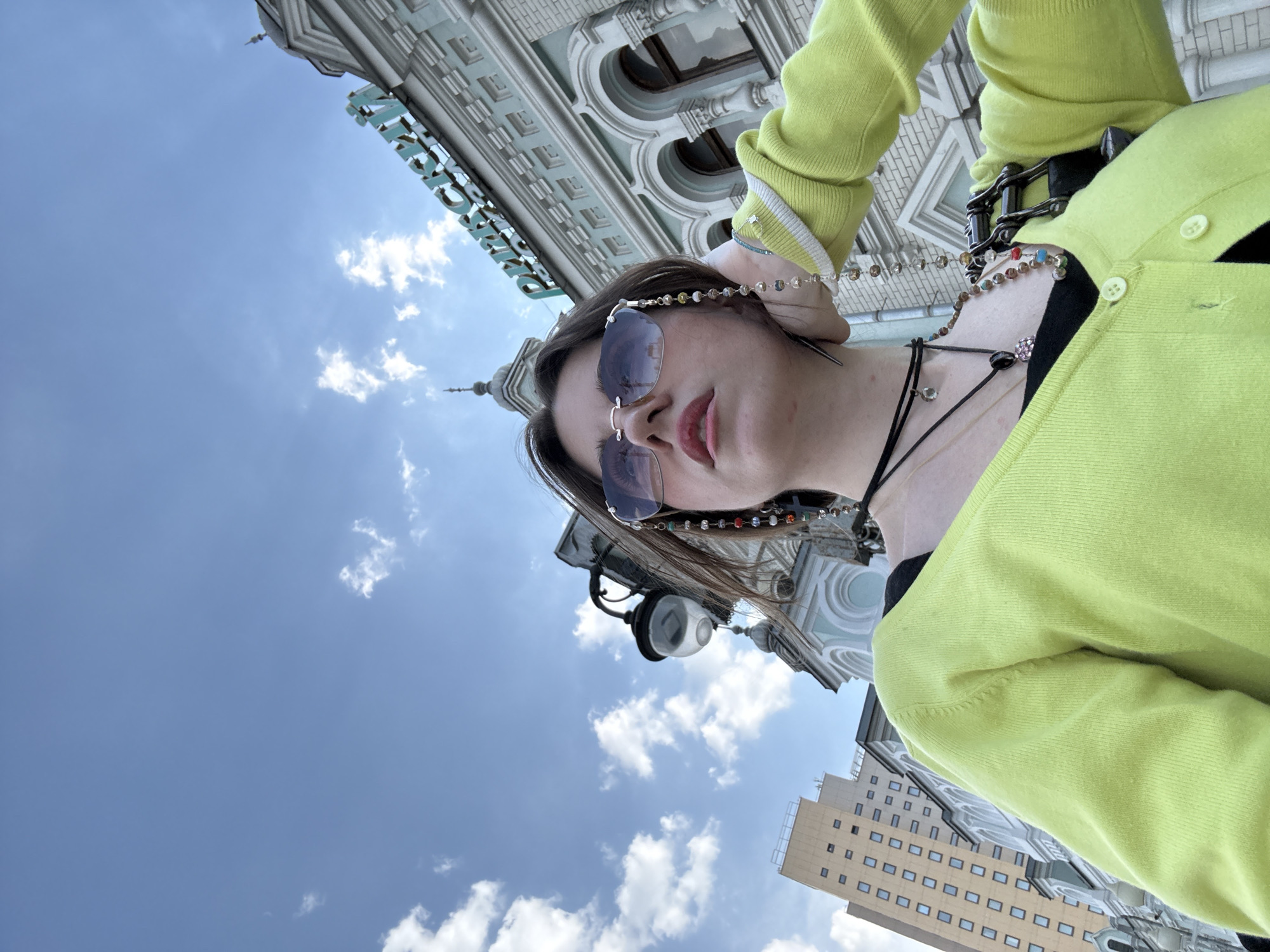 A low-angle portrait shows a person in a lime green sweater, sunglasses, and multiple necklaces, against a backdrop of buildings and a cloudy sky.
The building in the background is the Detsky Mir (Children's World) department store, located in Moscow, Russia. The building has a light-colored facade with ornate architectural details, including arches and columns. A modern building is partially visible at the lower right corner of the frame. The sky is blue with scattered clouds. A street light with a white orb is also visible.