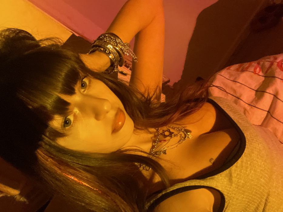 A woman lies on a striped bed under warm, yellowish lighting. She has bangs and dark hair with a glittery accent strand. Multiple silver bracelets adorn her raised arm. She wears a gray tank top with black trim and two necklaces, one with a Star of David. A small heart tattoo is visible on her chest. She looks up at the camera with a slight smile.