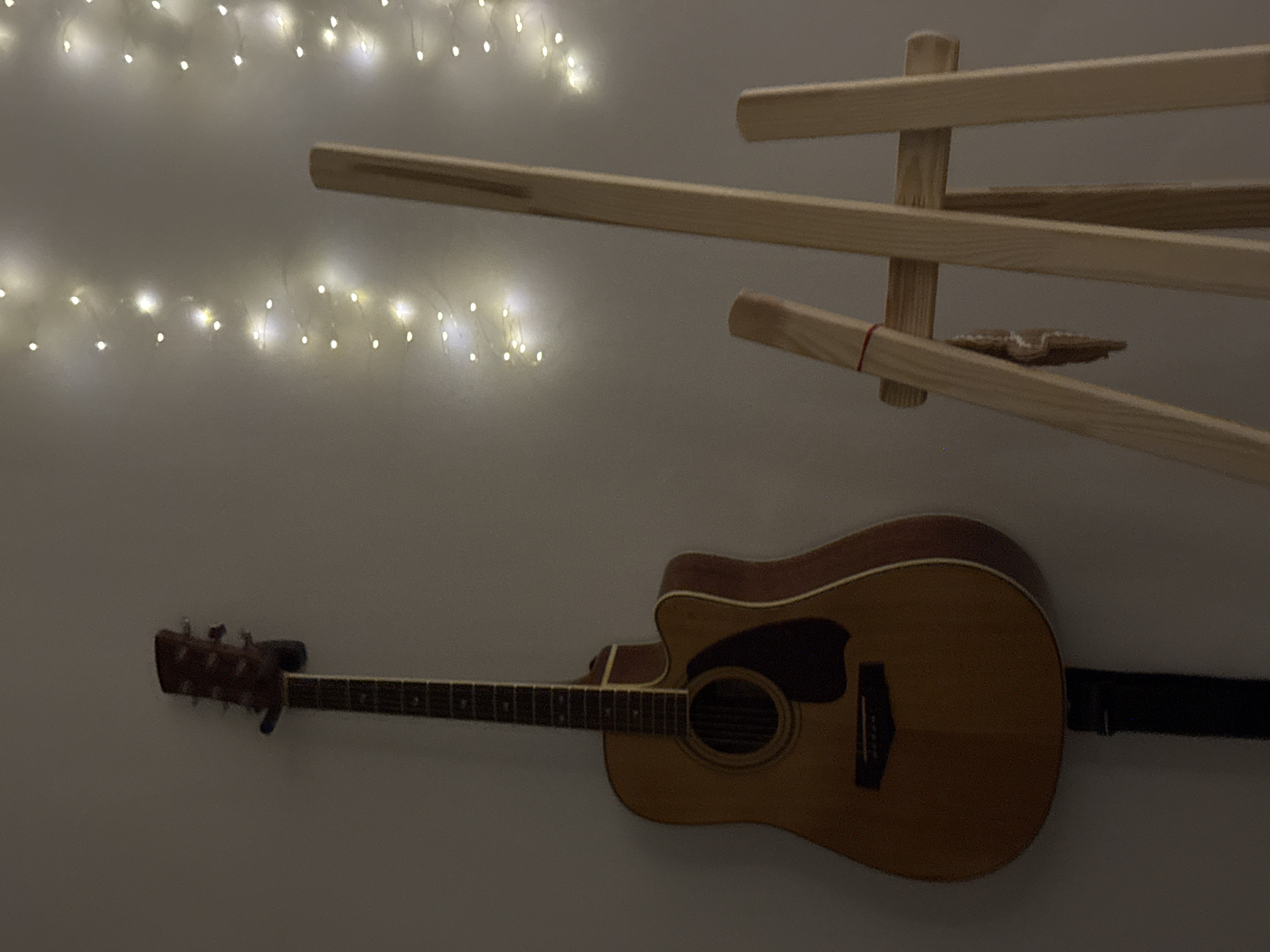 An acoustic guitar with a cutaway body and a dark pickguard is mounted on a wall with a black wall mount. To the right of the guitar is a wooden easel. The background is a plain wall with rows of small, warm-toned fairy lights. The lighting is dim, creating a soft ambiance. The scene appears to be indoors, likely in a domestic setting. There are no people visible in the image.