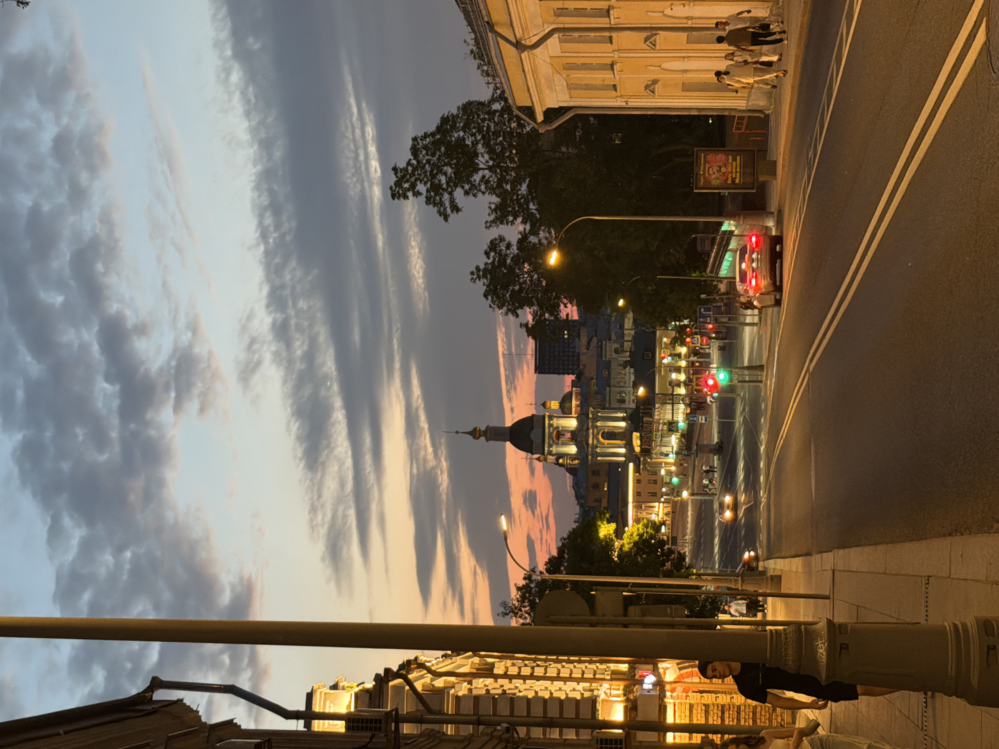 The image depicts a street scene in Moscow, Russia at dusk. A young man in a black t-shirt and shorts stands on the sidewalk, partially obscured by a lamppost. Several other pedestrians, dressed in casual attire, walk along the sidewalk on the right side of the street. Cars are visible on the road, some with their headlights on. Traffic lights are illuminated, with one showing a green light. In the background, a large building with a dome and multiple windows, illuminated by artificial lights, dominates the skyline. The sky is a blend of blue and orange hues, with scattered clouds. The architecture suggests a historic part of the city.