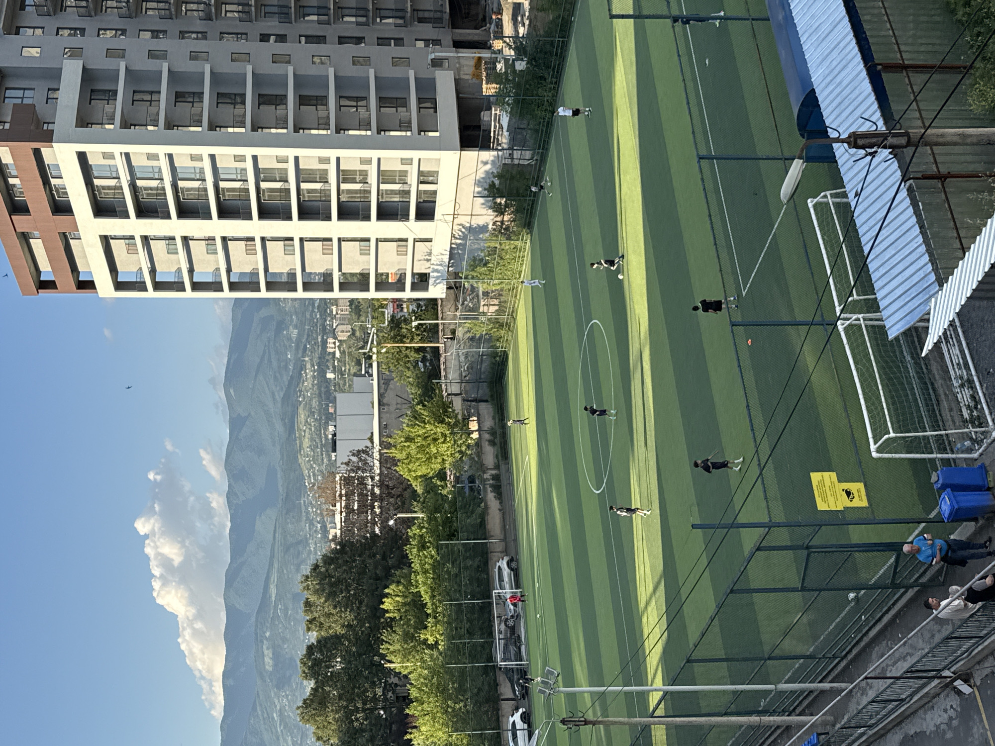 In Tbilisi, Georgia, an outdoor football field is the scene for a casual game. Several young men, dressed in athletic attire, are spread across the green turf, some actively participating in the match, while others stand ready. The field is bordered by a dark green fence, with a modern, multi-story residential building visible in the background, its windows reflecting the sky. Beyond the building, rolling, tree-covered hills rise to meet a hazy blue sky, where a few wispy clouds are present. The sun casts long shadows across the field, indicating it is late afternoon or early evening.