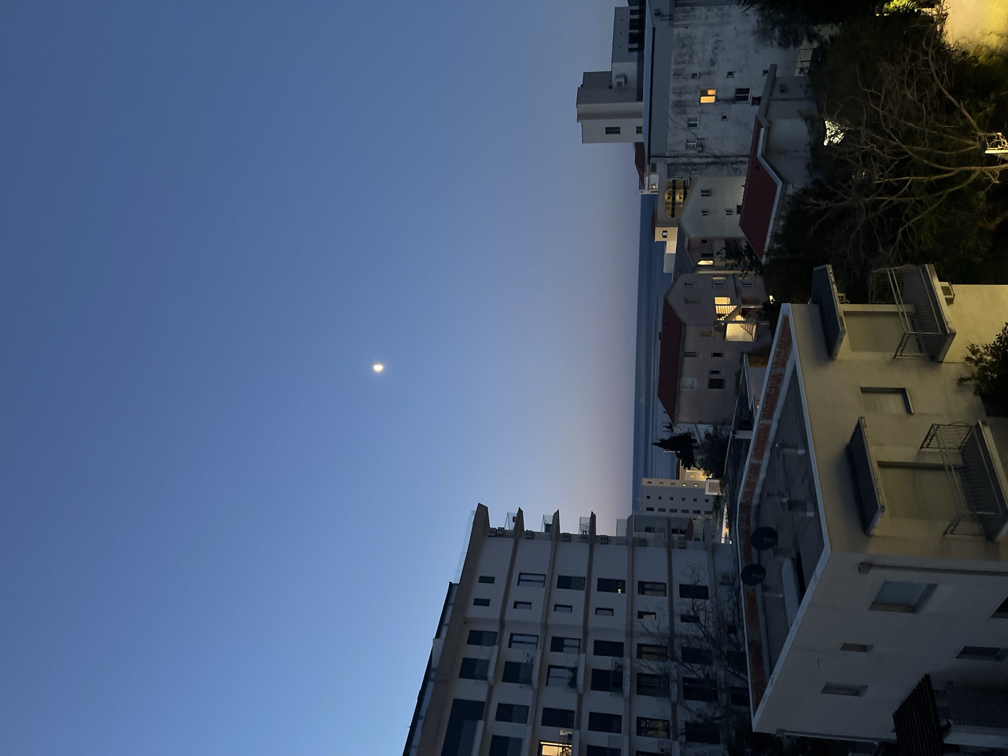 The image depicts a nighttime cityscape in Boreti, Montenegro. The sky transitions from a dark blue at the top of the frame to a lighter blue/purple horizon. A small, bright moon is visible in the upper left portion of the frame. Buildings of varying heights and architectural styles occupy the right side and bottom of the frame. The buildings are mostly white or light-colored, with some having red-tiled roofs. Some windows are lit, indicating occupancy. Trees and other vegetation are interspersed among the buildings. The perspective suggests the photograph was taken from an elevated position. The overall ambiance is calm and peaceful. A body of water is present between buildings near the horizon line. The scene is devoid of people.