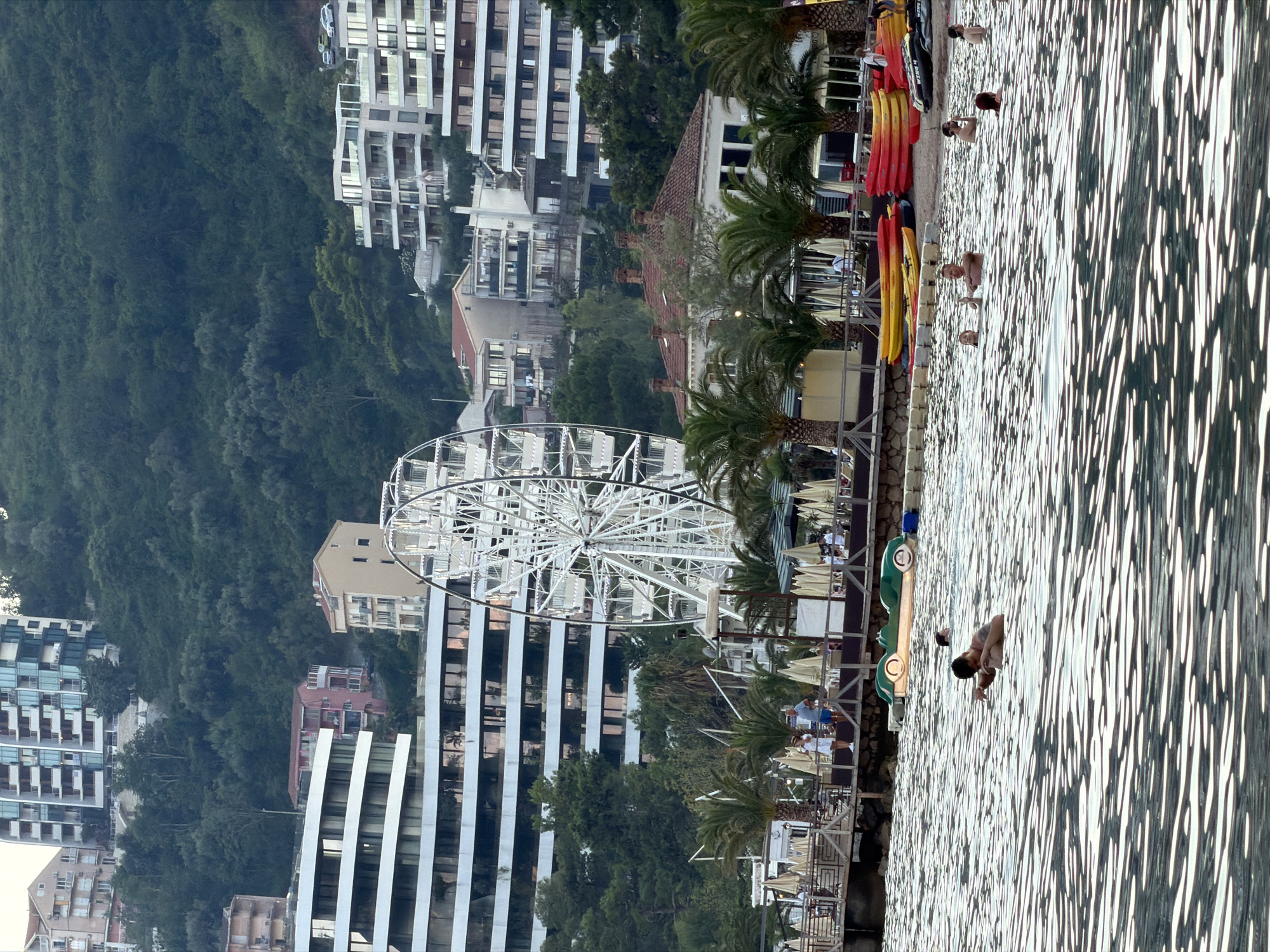 The image depicts a coastal scene in Budva, Montenegro. In the foreground, the sea is filled with rippling sunlight reflections, and several people are swimming. In the mid-ground, a wooden pier with umbrellas and palm trees lines the shore. Behind the pier, a white Ferris wheel stands prominently, with buildings and a tree-covered hillside rising in the background. Several colorful kayaks and pedal boats are visible on the beach and in the water. The buildings in the background appear to be hotels or apartment complexes. The overall impression is a lively tourist resort area.