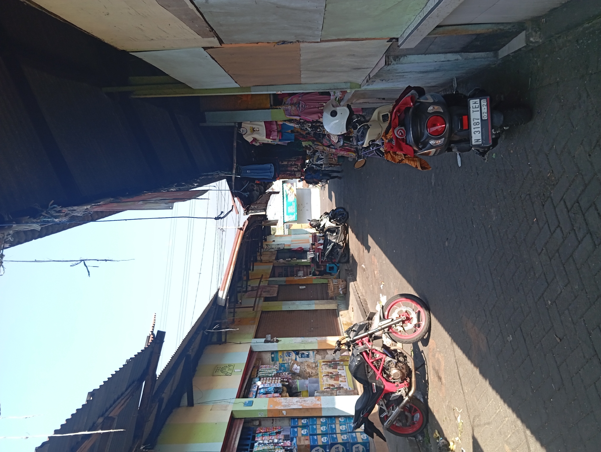 The image shows a narrow street in a marketplace in Kepulungan, Indonesia, lined with shops on both sides. The street is paved with brick. Two motorcycles are parked in the foreground. The one on the left is a red and black sportbike. The one on the right is a white scooter. Several people are walking in the background. The shops appear to sell clothing and general goods. The sky is clear and blue.