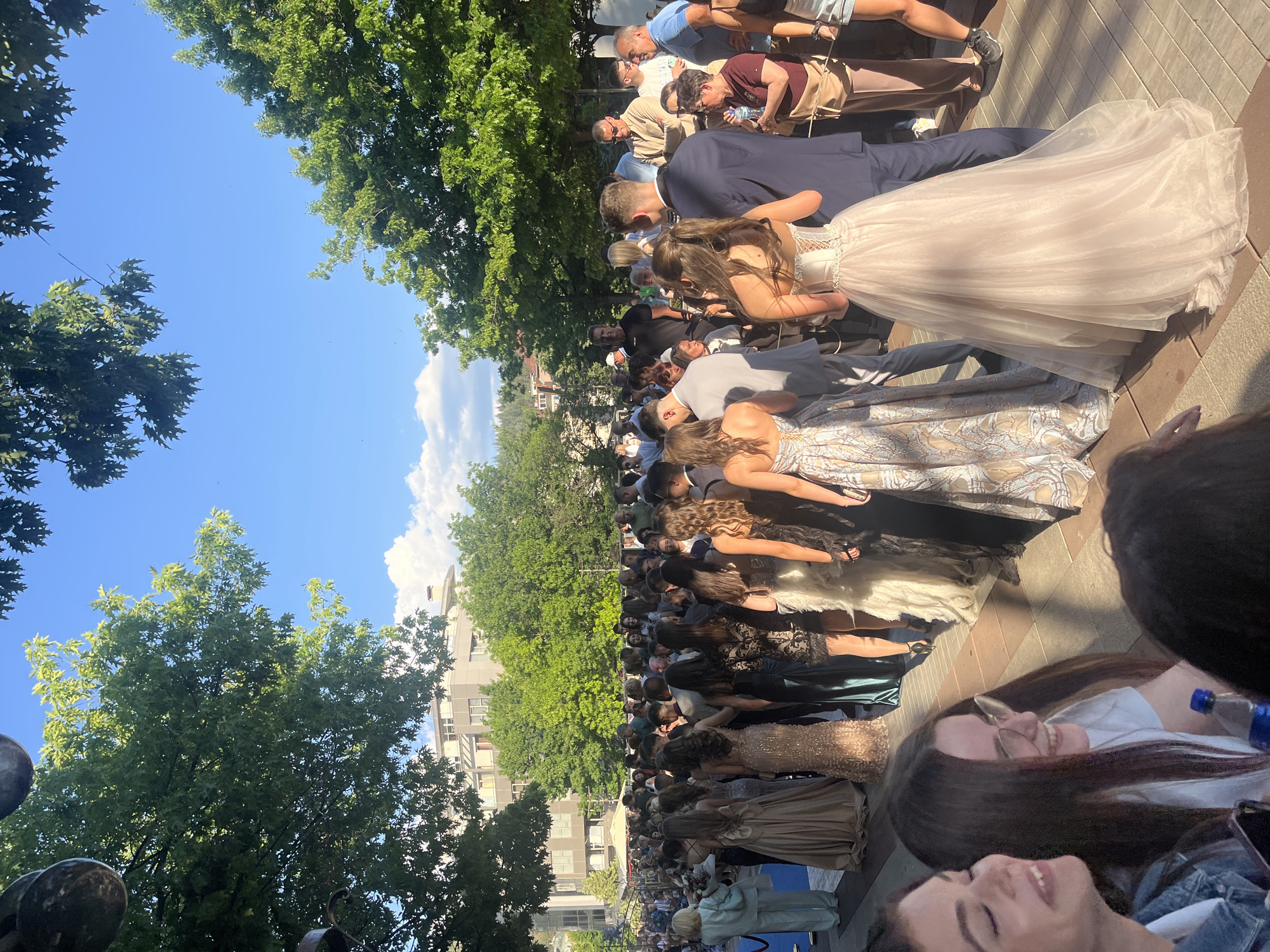 The image depicts a graduation ceremony in Blagoevgrad, Bulgaria. A large group of graduates, mostly in formal dresses, is lined up on a paved area next to a street with buildings and trees. The sky is blue with some clouds. A couple is walking in the foreground. Spectators and families are standing near the trees. The focus is on the graduating class.