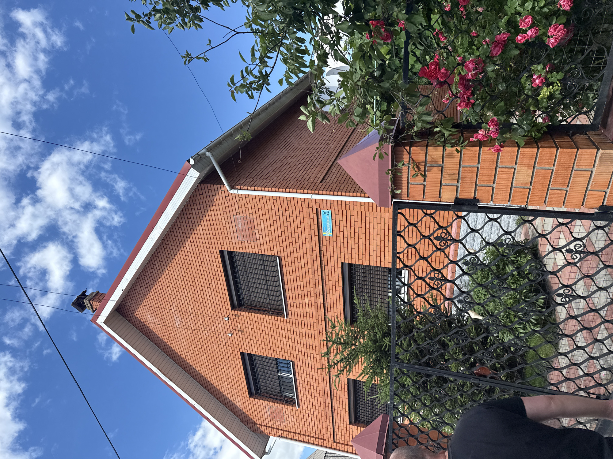 The image shows a brick house with a red tiled roof, surrounded by greenery. A black metal fence with decorative scrollwork encloses the property, and a flowering bush with pink roses is visible on the right side of the frame, climbing over the fence. There are several windows on the house, all fitted with metal security bars. A street sign with Cyrillic text and the number "20" is attached to the brick wall of the house. The sky is blue with scattered white clouds, suggesting it is daytime. The overall impression is one of a residential property, possibly in a suburban or rural setting in Obukhivka, Ukraine, given the visual cues. The scene appears peaceful and serene, with no specific activity taking place.