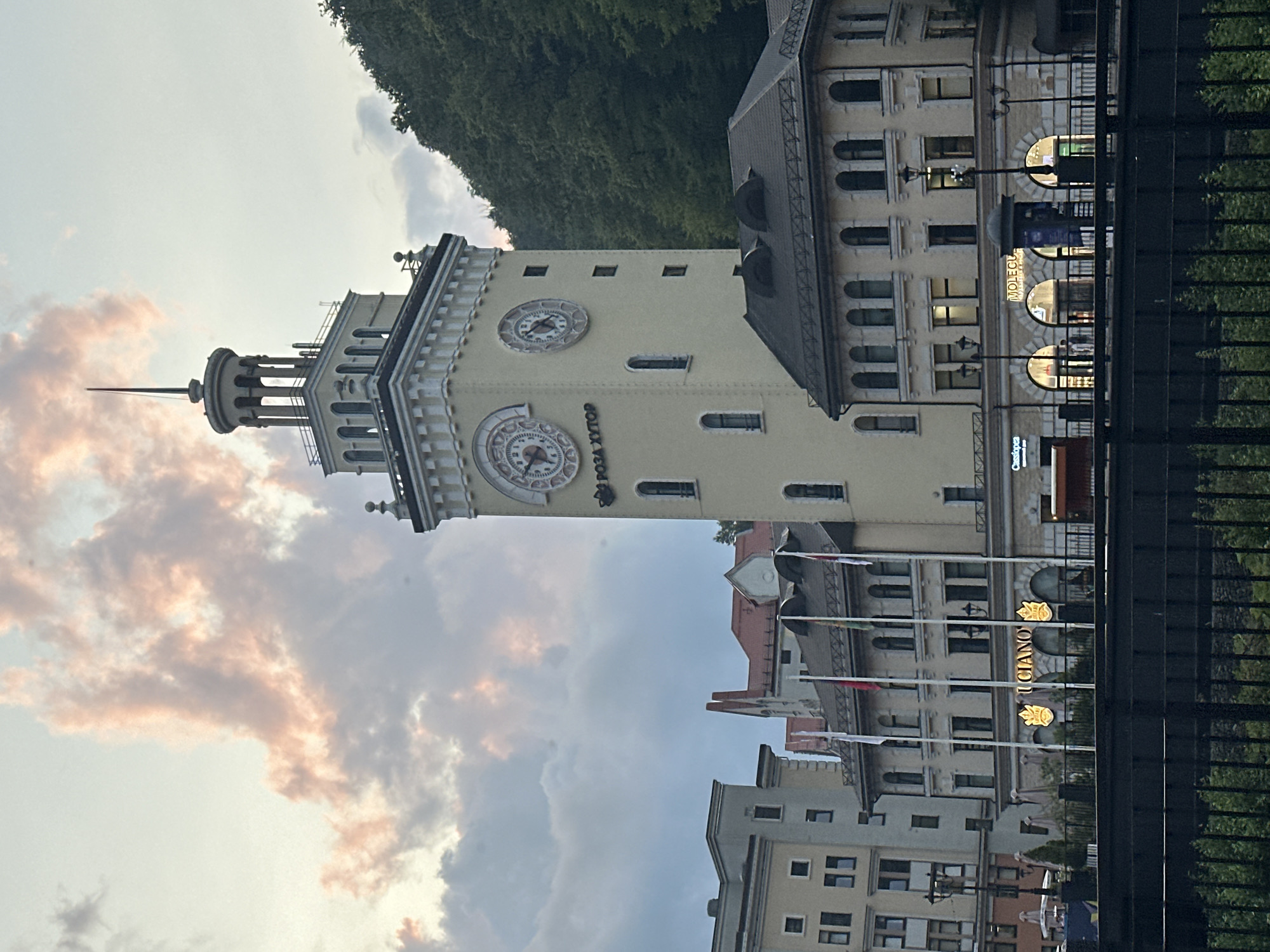 An outdoor, eye-level, medium shot captures the Rosa Khutor Clock Tower in Estosadok, Russia, bathed in the soft light of a dusk sky filled with dramatic, pink-tinged clouds. The tower, a prominent structure, stands tall with a clock face visible, and a spire crowning its top. To the left of the tower, modern-style buildings with visible windows line the street. Flags fly from poles in front of a building to the left of the clock tower. The scene is framed by a dark, horizontal fence in the foreground, with greenery visible beneath it. The background features a densely forested hillside.