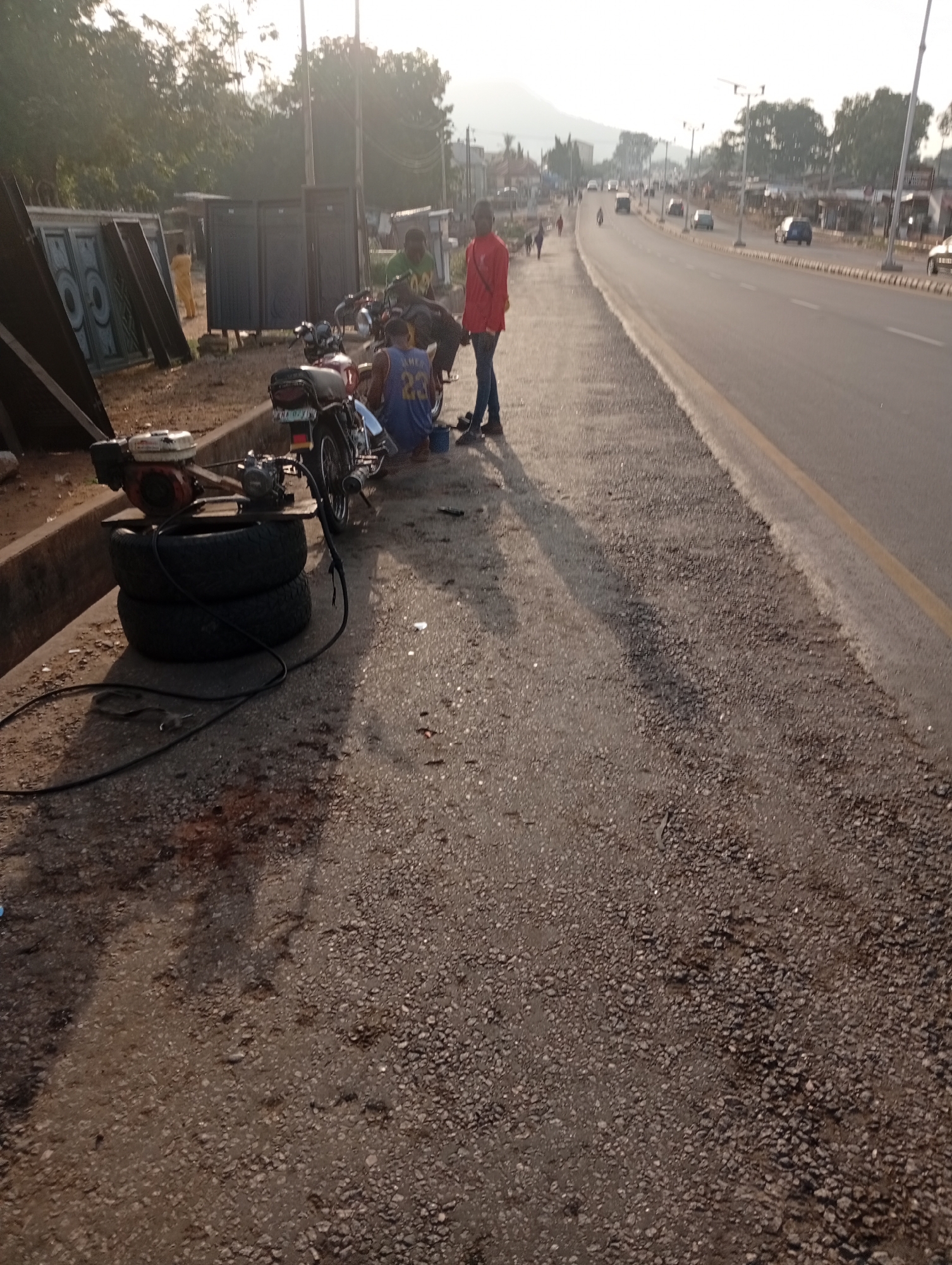 The image depicts a roadside scene in Adankolo, Nigeria. A group of people are gathered on the side of a paved road, near a motorcycle repair setup. Two motorcycles are visible. A small engine sits on top of two stacked tires, with wires extending to the ground. Three men are present. One man stands wearing a red shirt, another man is wearing a blue tank top with the number "23" on it, and another man is wearing a green shirt. A pile of metal doors lean against a concrete divider. In the background, a paved road with multiple vehicles is visible. Trees and a distant mountain can be seen in the hazy background.