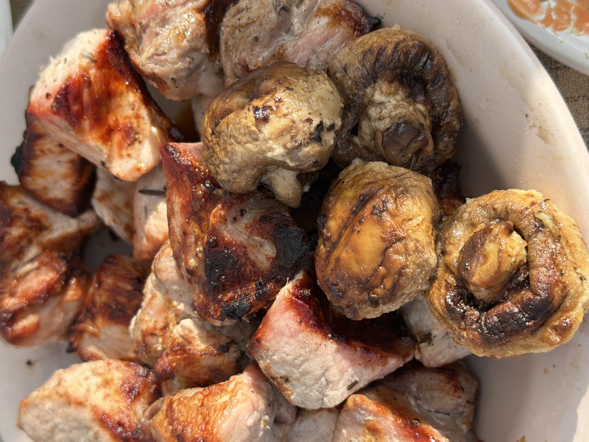 The image shows a close-up of a plate of grilled food, likely shashlik, consisting of pieces of meat and whole mushrooms. The meat is cut into bite-sized cubes and has a browned, slightly charred exterior with visible grill marks. The mushrooms are also grilled and appear to be button mushrooms, some with their stems attached. The food is arranged on a light-colored, possibly ceramic, plate. The lighting suggests an outdoor setting, possibly during daylight hours, with shadows indicating a sunny environment. The background is slightly out of focus, but hints of a table surface covered with a textured cloth can be seen. The overall impression is of a freshly prepared meal, ready to be served or eaten. No people are visible in the image. The specific location in the city of Chaikovsky, Russia cannot be determined from the visual information provided.