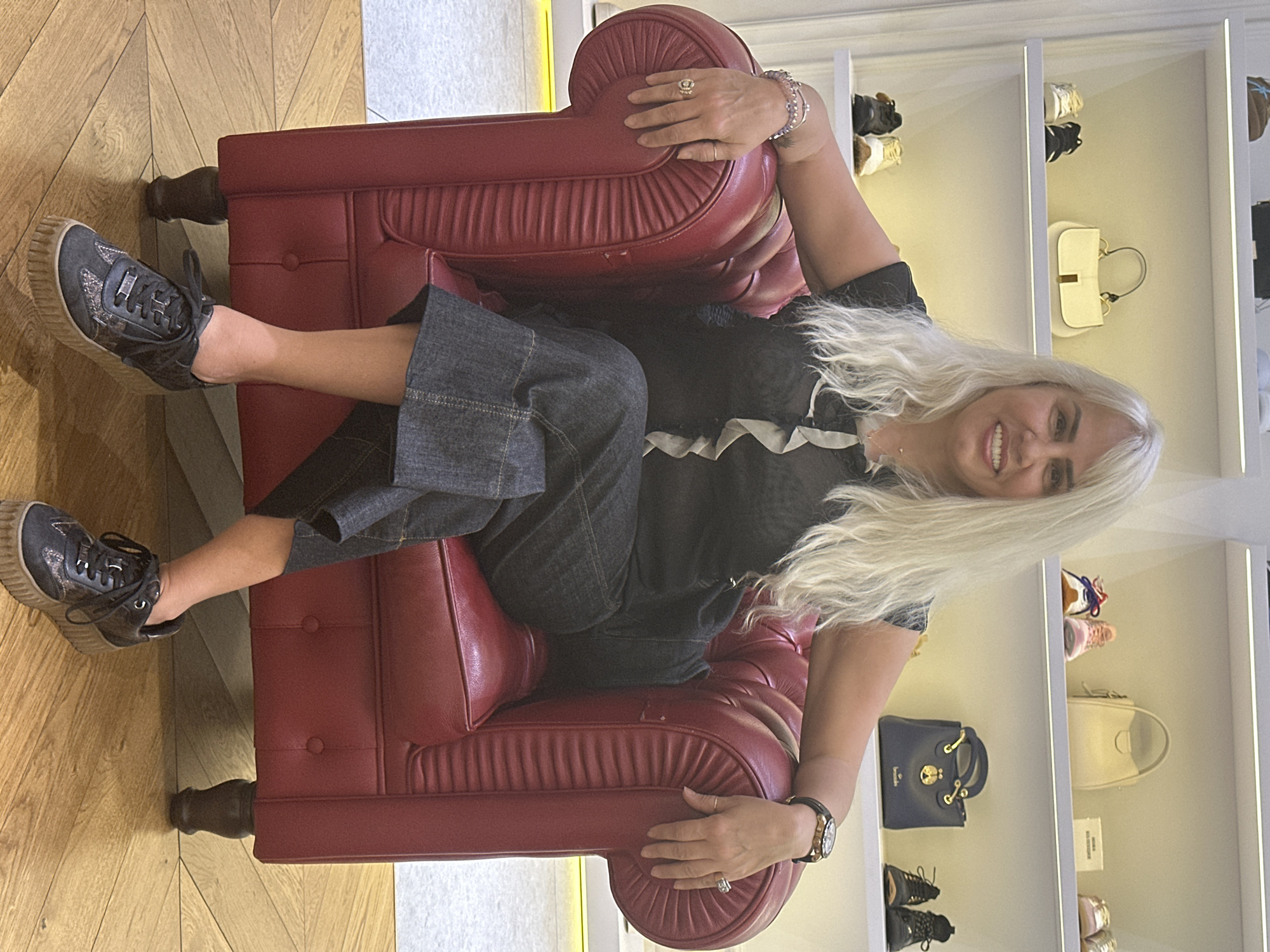 A woman with long platinum blonde hair is reclining in a red leather armchair. She is wearing a black top with a white ruffled collar, dark denim culottes, and black sneakers with thick, tan-colored soles. She has multiple rings on her fingers and a watch on her left wrist. Her arms are resting on the arms of the chair.
Behind her is a white shelving unit with built-in lighting, displaying shoes and handbags. The background is softly lit, suggesting a well-lit interior space. The presence of shoes and handbags suggests a retail environment, possibly a high-end boutique. The floor is covered in light-colored hardwood.
There is no evidence to determine that the picture was taken in Sofia, Bulgaria.