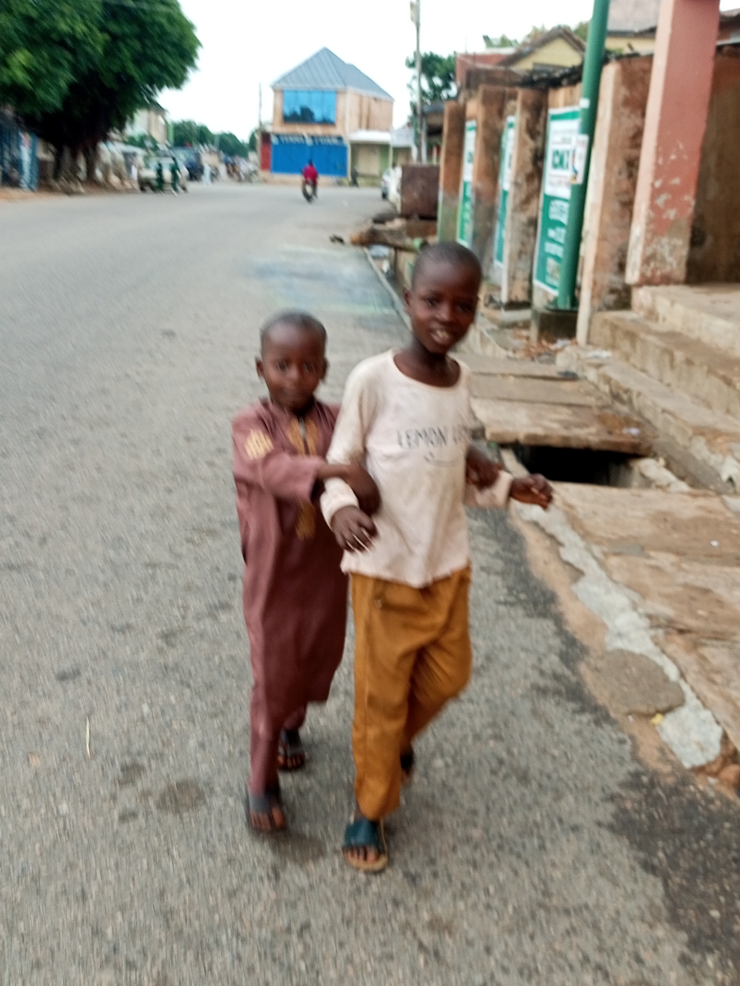 Two young boys are walking on an asphalt road in Gombe, Nigeria. The boy on the left is wearing a brown garment and the boy on the right is wearing a white t-shirt with text and brown pants. They are walking towards the viewer. The road is lined with buildings and a tree on the left side of the image. There is also a storm drain on the right side of the image. The scene appears to be in an urban or semi-urban area. The sky is overcast.
