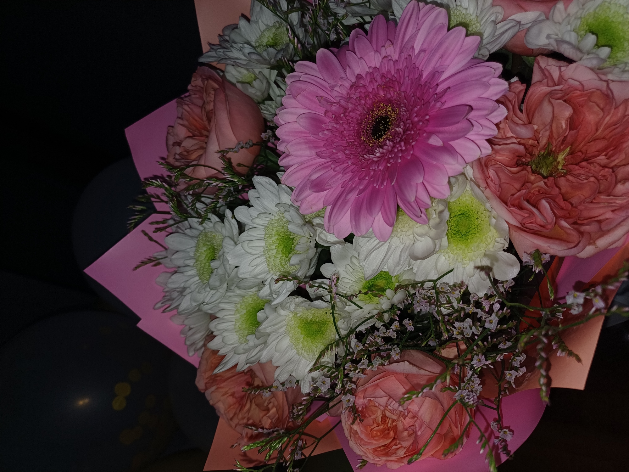 This image features a close-up view of a vibrant bouquet of flowers. The primary subjects are the flowers themselves, which include a large pink gerbera daisy, several white chrysanthemums with green centers, and several peachy-pink roses with intricate petal formations. Delicate sprigs of small, light purple flowers and green foliage are interspersed throughout the bouquet. The flowers are arranged in a way that suggests they are presented as a gift, possibly in a bouquet wrapper that is partially visible in shades of pink and peach.
The background is dark and out of focus, with hints of what appear to be dark balloons and some faint, out-of-focus yellow lights on the left side, suggesting a celebratory or festive occasion, perhaps an indoor event or evening setting. The lighting appears to be artificial and somewhat dim, casting shadows that add depth and highlight the textures of the flowers. There is no visible text in the image. The overall mood conveyed by the image is one of beauty and celebration, with the rich colors of the flowers being the main focus. The composition is from a top-down perspective, looking into the heart of the arrangement.