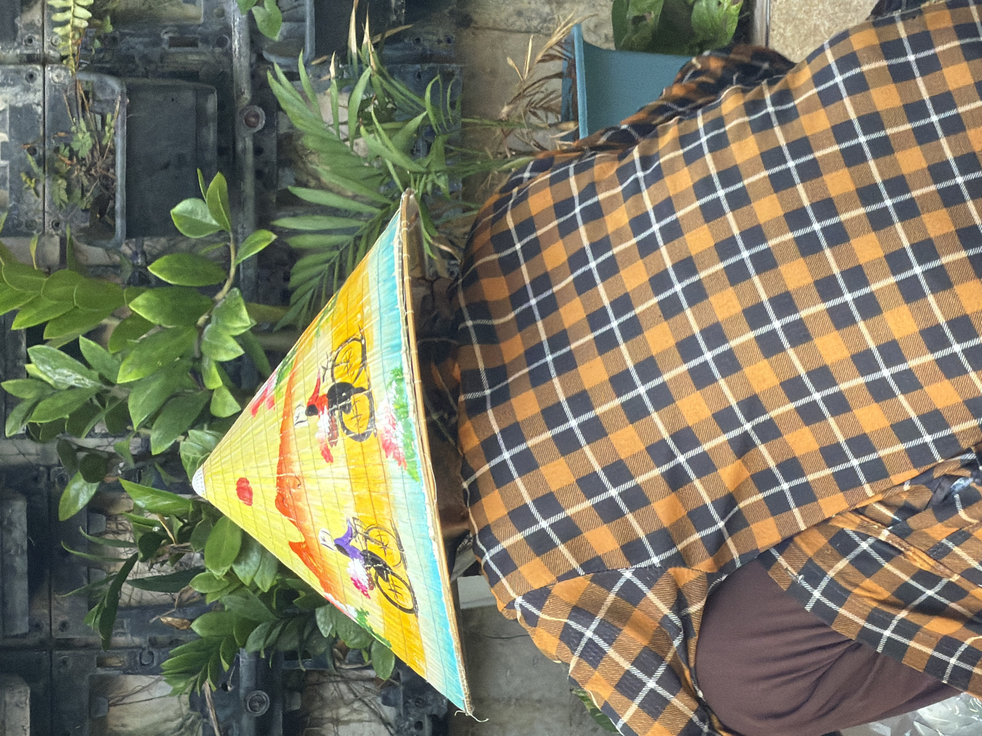 A person wearing a conical hat with a painted scene of people on bicycles and a plaid shirt is squatting in front of a wall with vertical planters and plants. The conical hat is yellow with red, blue, and white accents, and depicts two figures on bicycles with baskets of flowers. The shirt is orange and black plaid with white stripes. The background consists of a dark, textured wall and green foliage. The scene appears to be outdoors, possibly in a garden or courtyard, with the implication of a location in Nha Trang, Vietnam, due to the conical hat style.