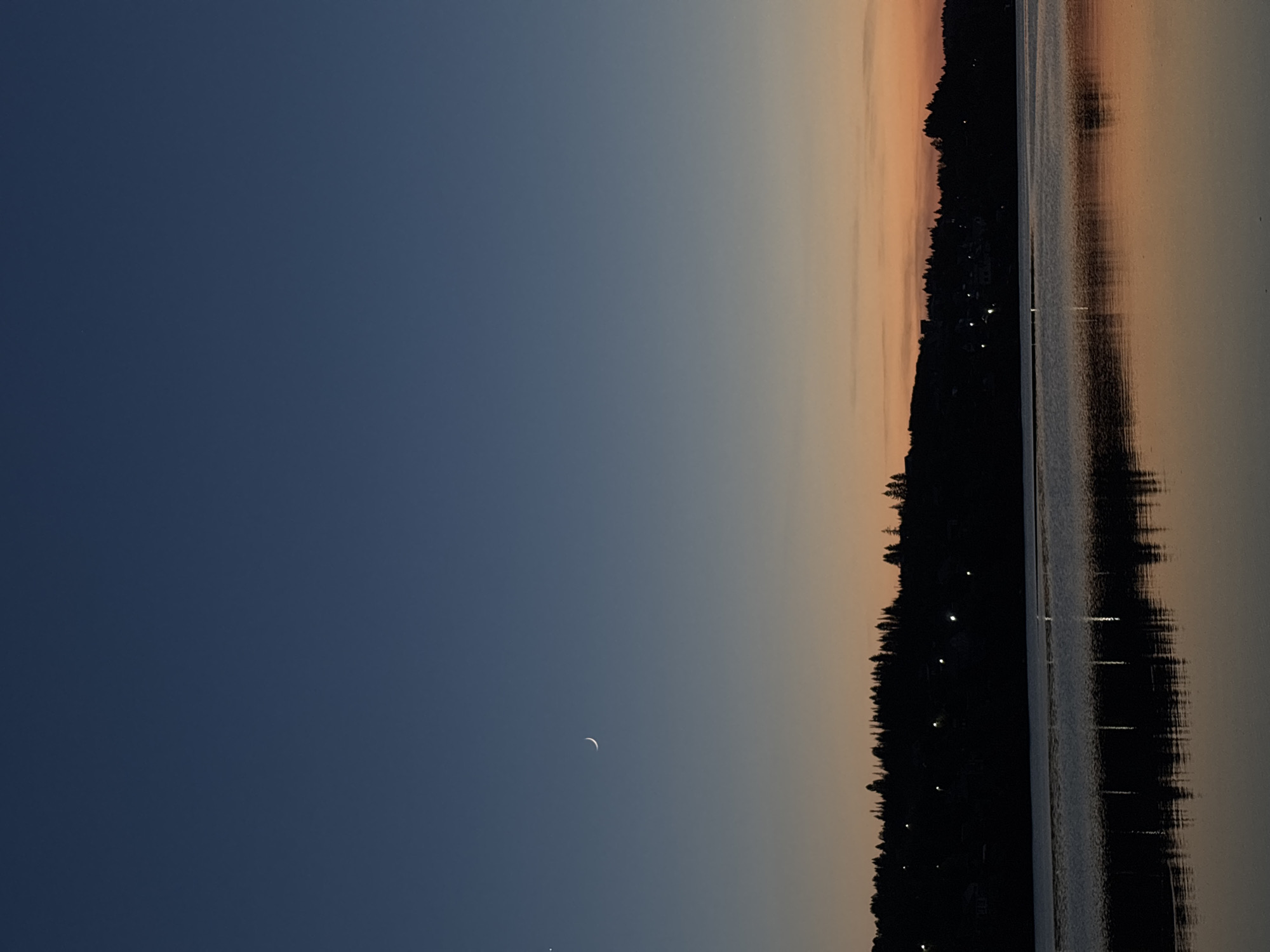 The image shows a twilight or early morning scene in what appears to be Gora (Khokhlovskoe S/P), Russia. A crescent moon is visible in the upper left portion of the image, set against a dark blue sky. The horizon is a blend of twilight colors, transitioning from a light orange hue near the skyline to a darker gradient towards the upper left. A line of trees, possibly marking the edge of a forest or shoreline, is visible along the horizon. Their silhouettes are dark against the fading light of the sky. To the right of the trees, there's a body of water with a reflective surface, capturing the colors of the sky and mirroring the tree line. Small lights, possibly from buildings, dot the silhouette of the tree line. The overall scene depicts a peaceful, natural setting at dusk or dawn.