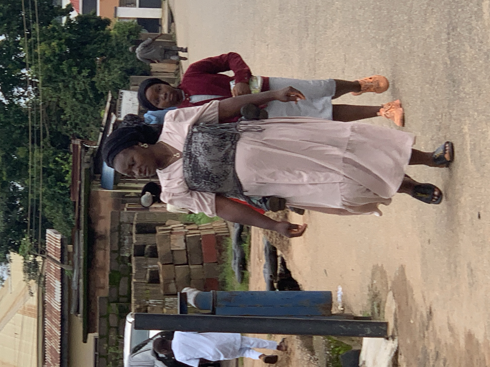 In Jos, Nigeria, on an outdoor paved street, a group of people are captured in a candid moment. The central figure, a woman wearing a light pink dress and a black headscarf, walks towards the viewer's right. She is adorned with a necklace and earrings, and her dress features a patterned overlay on the chest. Beside her, a young girl in a maroon jacket and grey skirt follows, holding a clear plastic bag with a yellow label. Further in the background, a man in a white traditional outfit walks away from the camera, while another man in grey walks further down the street. The environment suggests an urban setting with buildings and a stack of bricks visible on the left side of the image, with lush green trees forming a backdrop. The lighting indicates an overcast day.